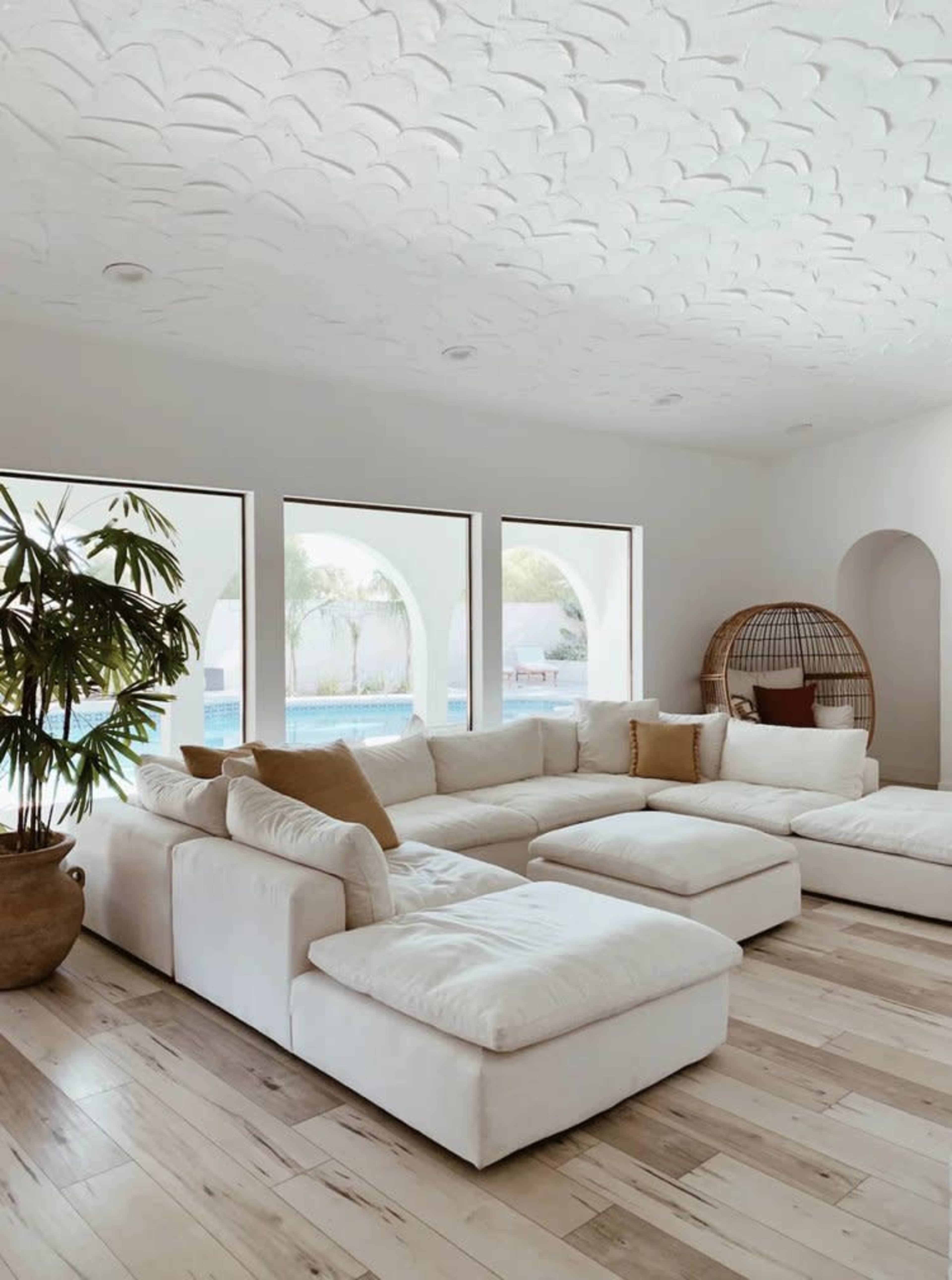 The image shows a light-filled living room with a large white sectional sofa, a decorative plant, and arched windows revealing a pool view.