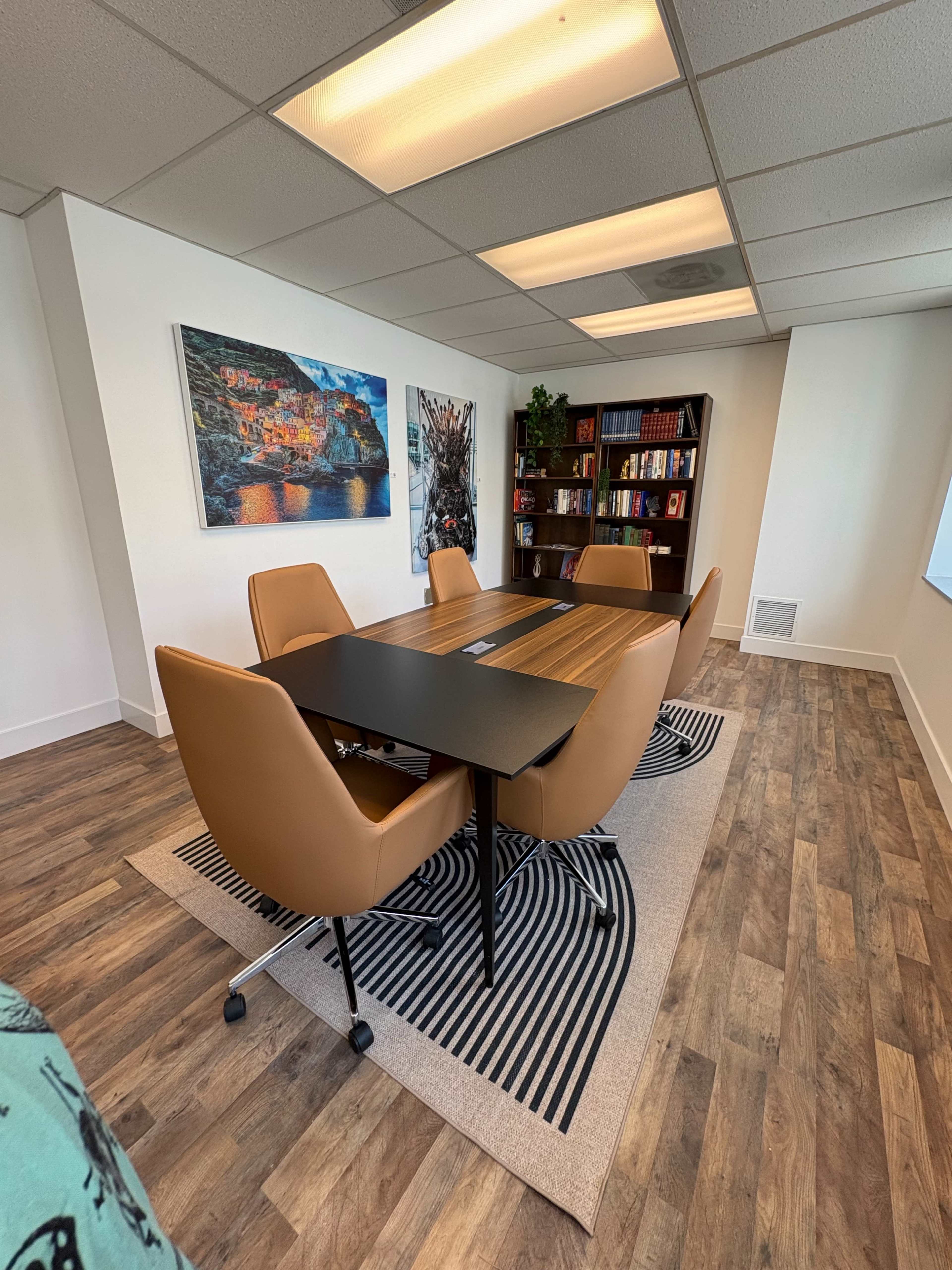 The image shows a modern conference room with a long table surrounded by four tan chairs, a bookshelf filled with books, and two paintings on the walls.