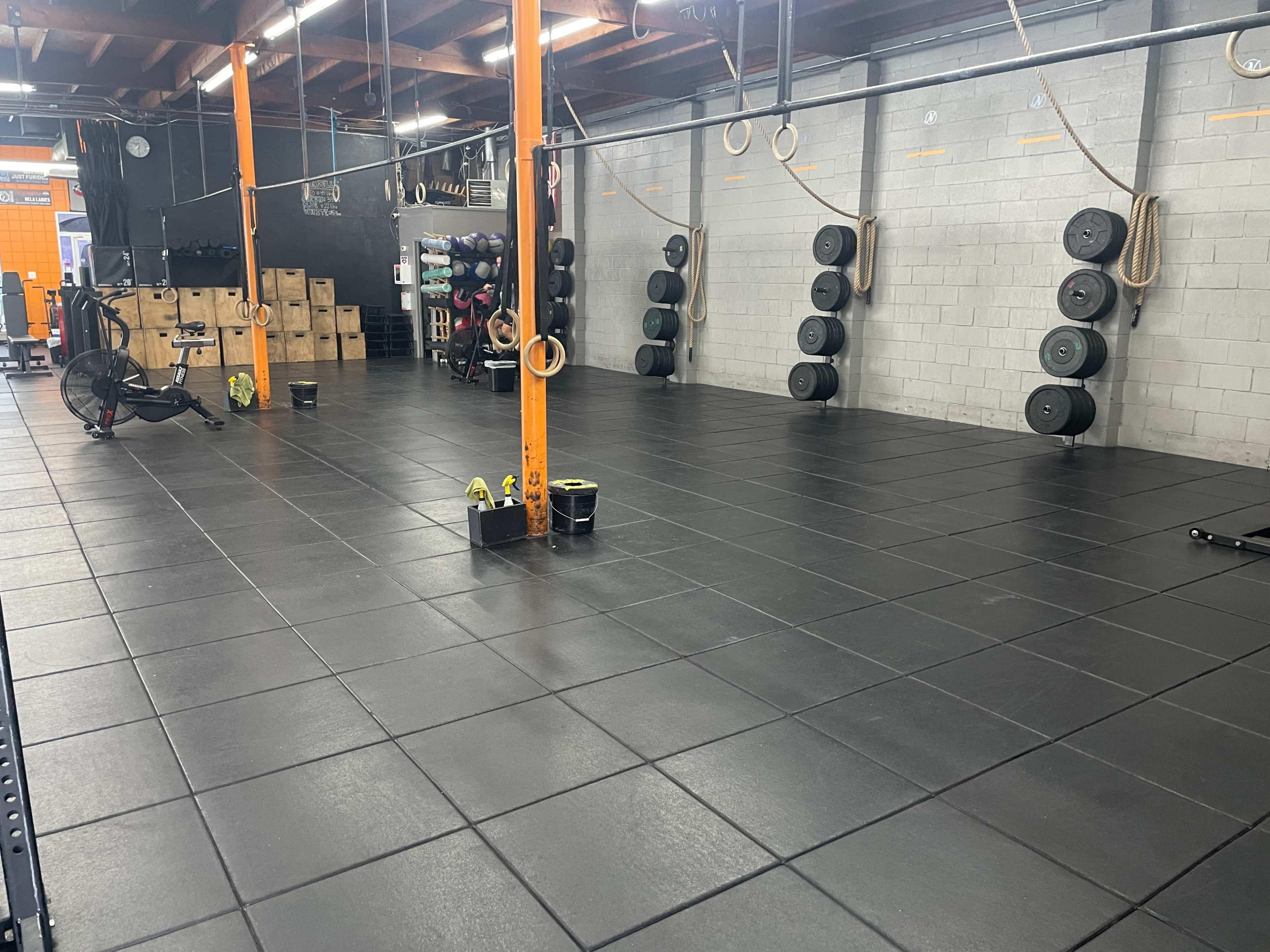 An empty gym with black rubber flooring, orange supports, and various exercise equipment along the walls.