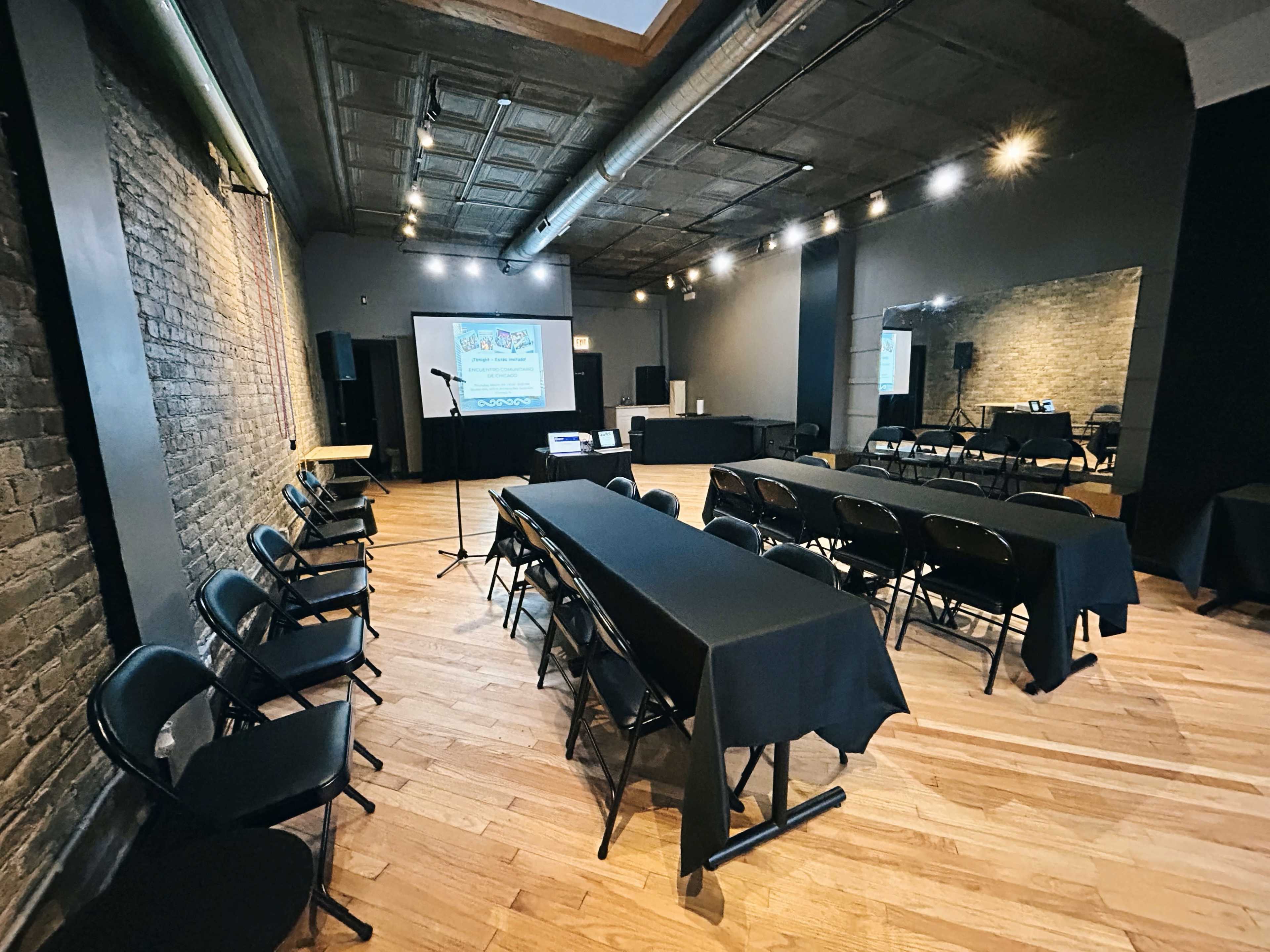 The image shows a seminar room featuring rows of black tables and chairs, a projector screen, and a microphone stand.