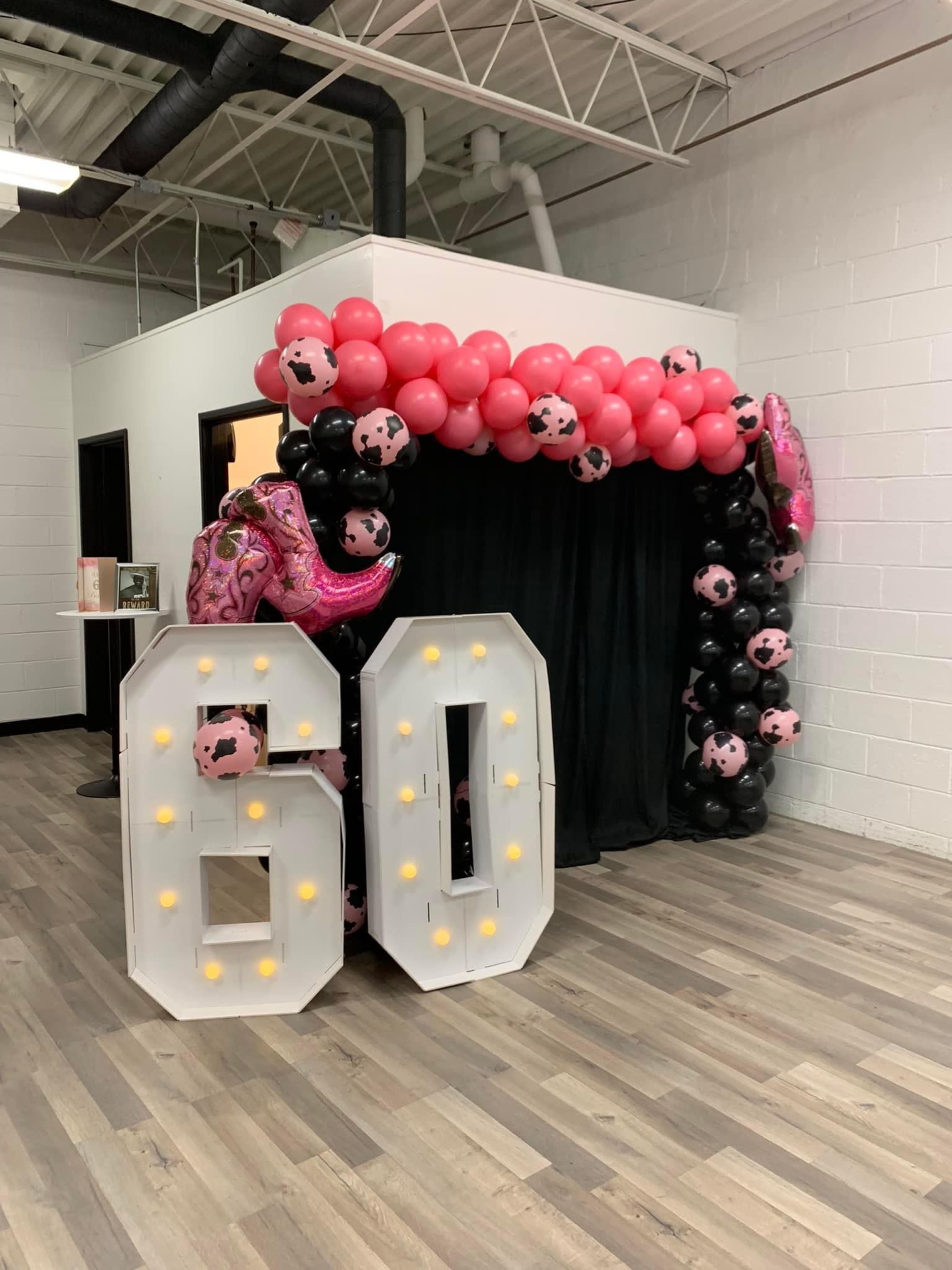 A large white "60" sign with lights stands beneath a decorative arch made of black, pink, and cow-print balloons.
