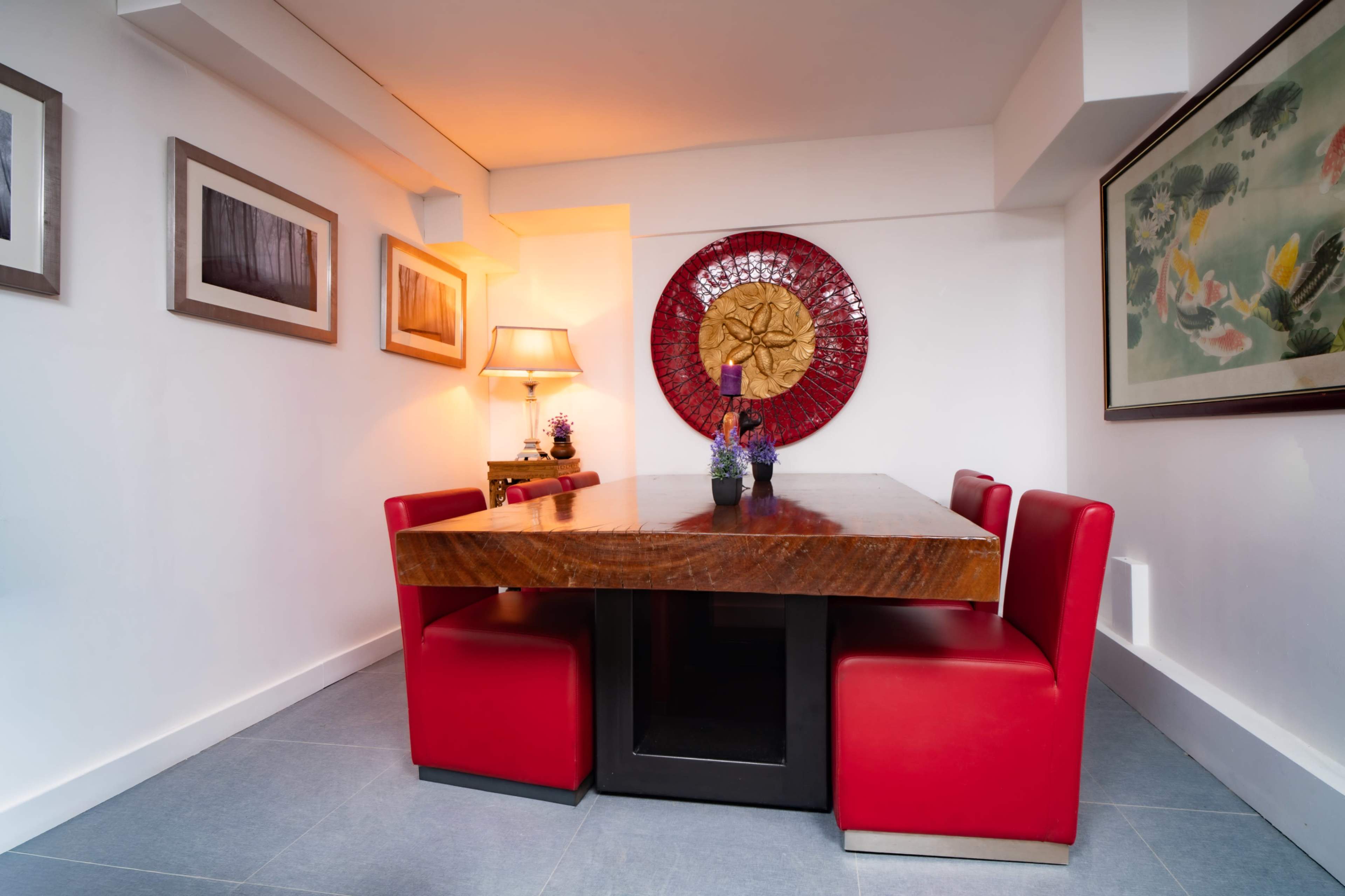 The image shows a dining area with a large wooden table surrounded by four red chairs, accented by a decorative wall piece and framed artwork.