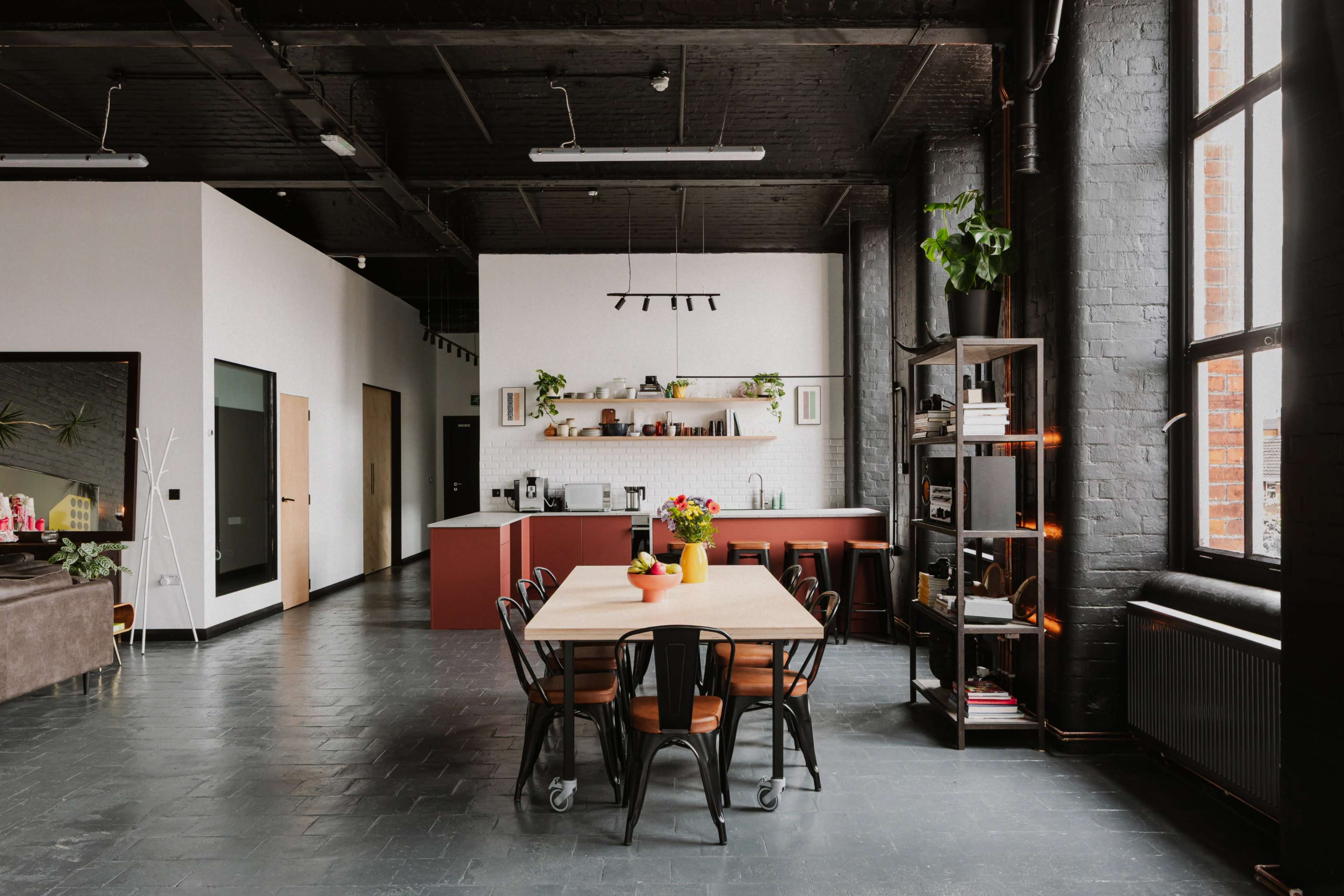 The image shows a spacious, industrial-style kitchen and dining area with a large wooden table surrounded by chairs, a modern kitchen with open shelving, and large windows.