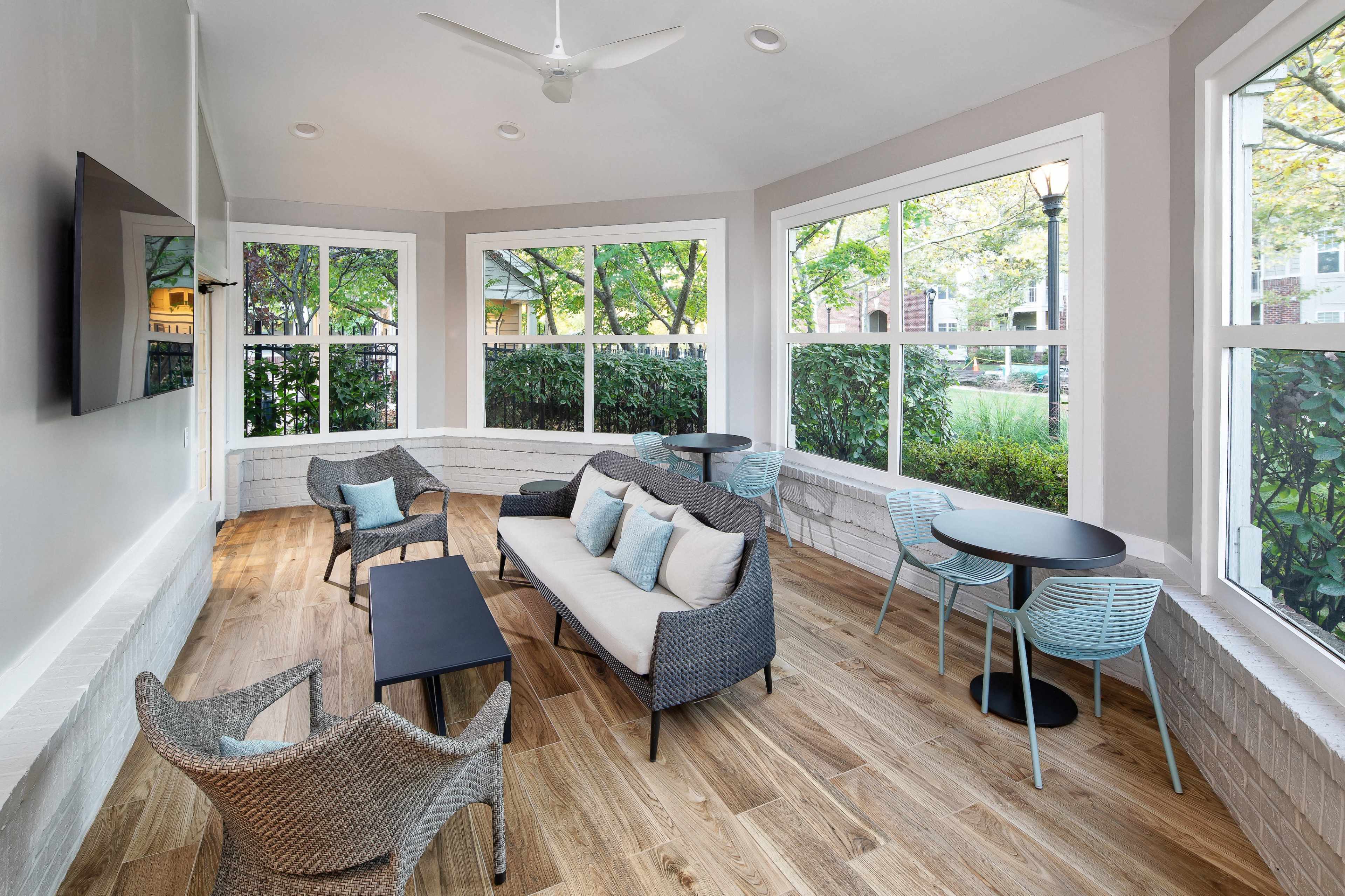 The image shows a sunlit lounge area with a sofa, two chairs, and tables surrounded by large windows and greenery.