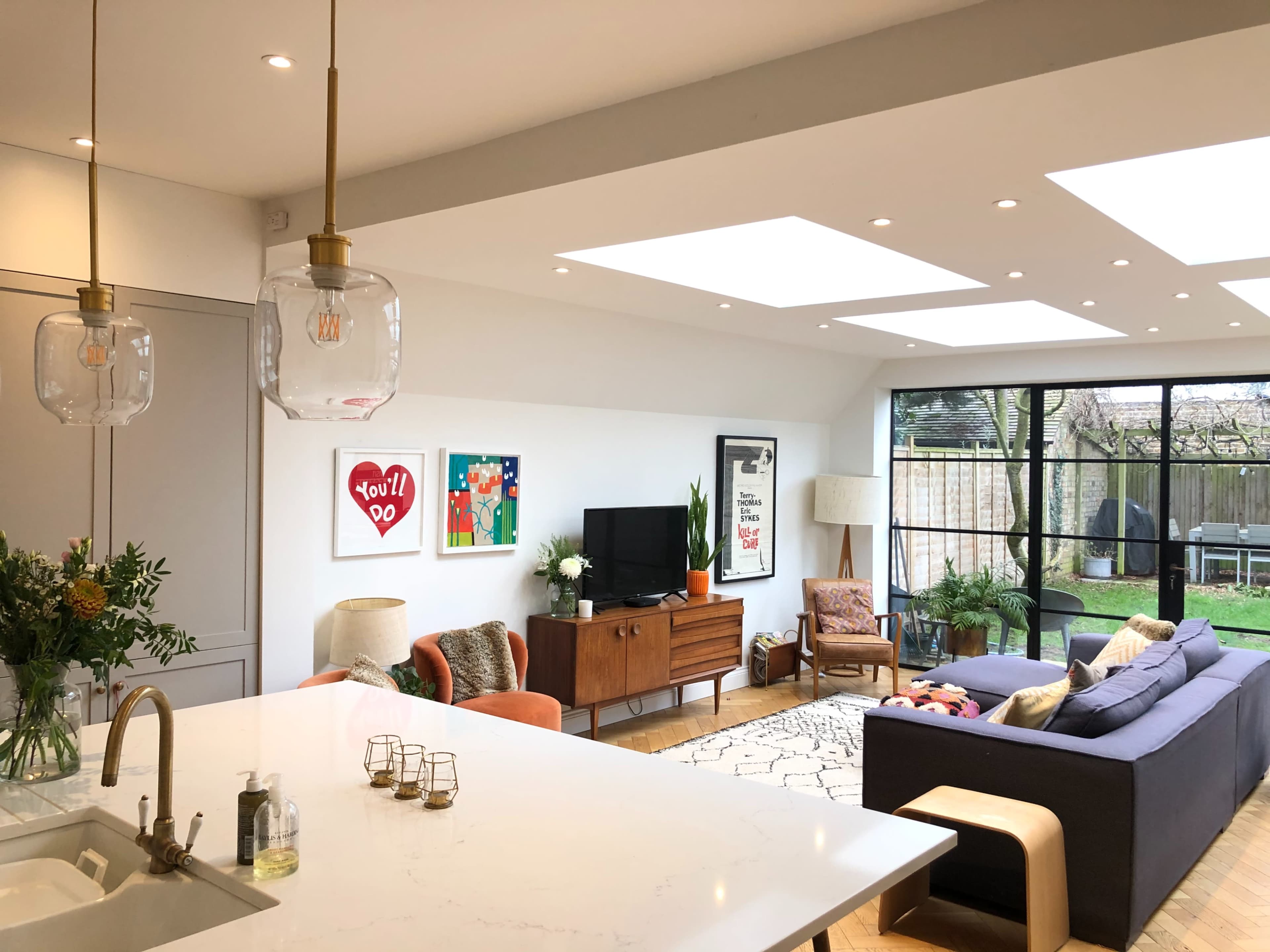 A modern living room features a large window, a comfortable sofa, a wooden sideboard with a TV, and a kitchen island with pendant lights.
