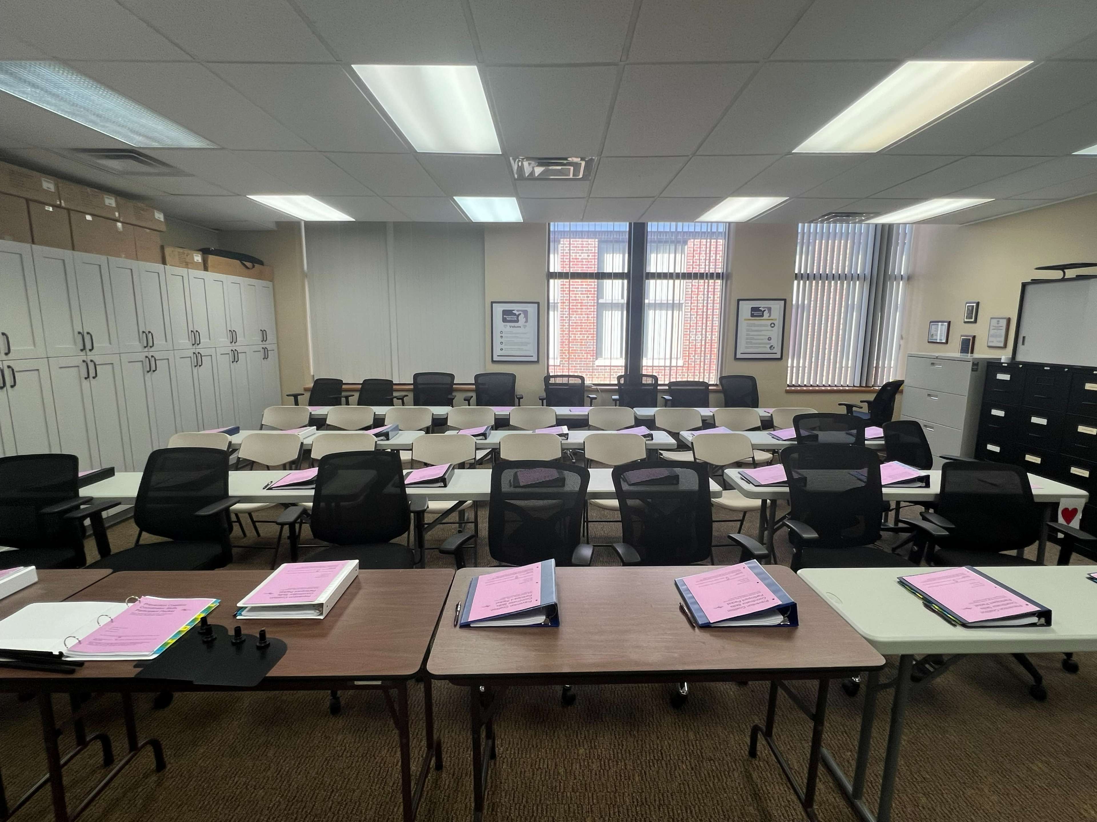 A classroom is organized with rows of tables and chairs, each decorated with pink folders, and storage cabinets line the walls.