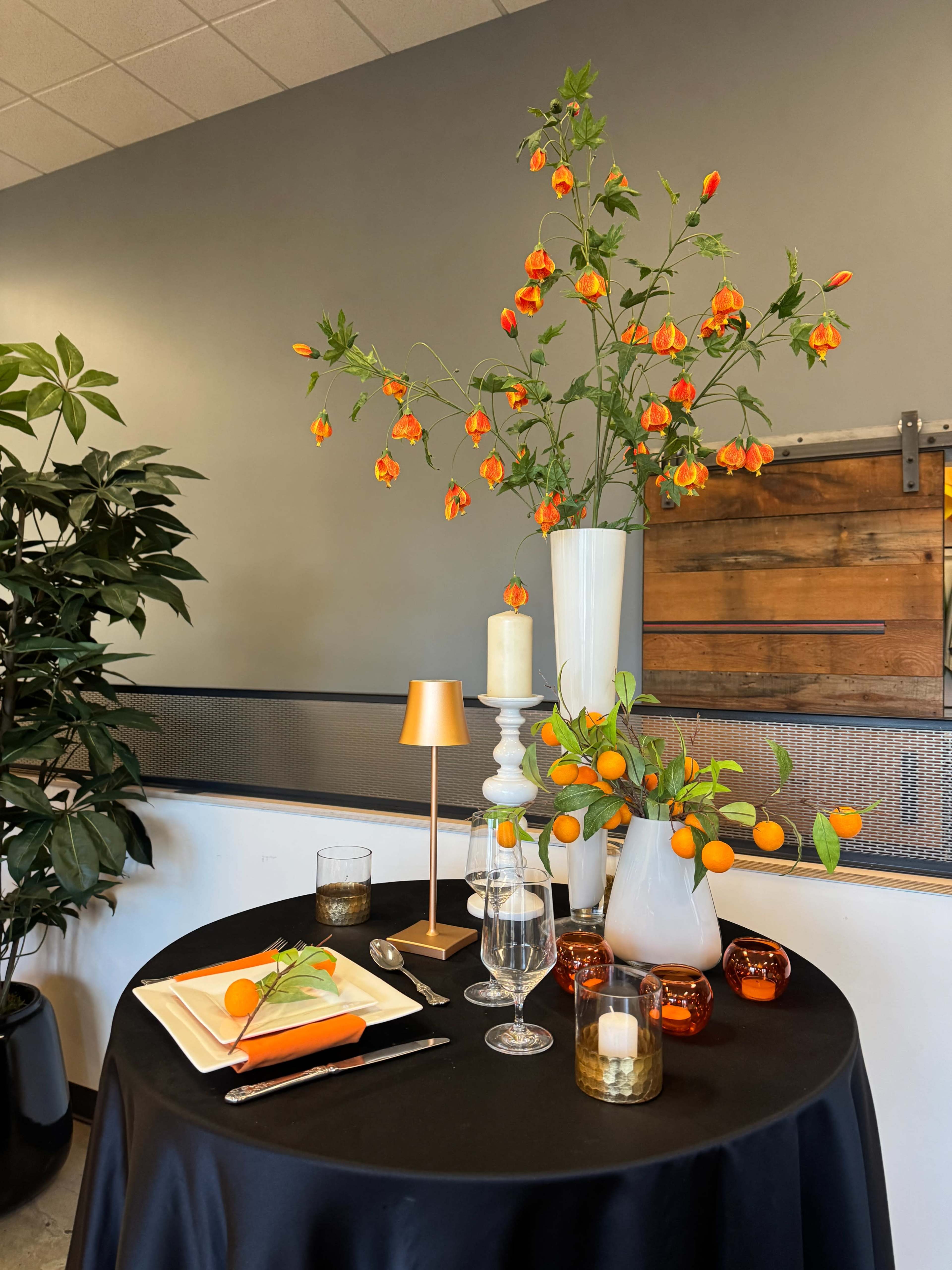 A round table is set with a black cloth, featuring decorative items like a white vase with orange blooms, candles, a glass of water, and a plate with oranges.