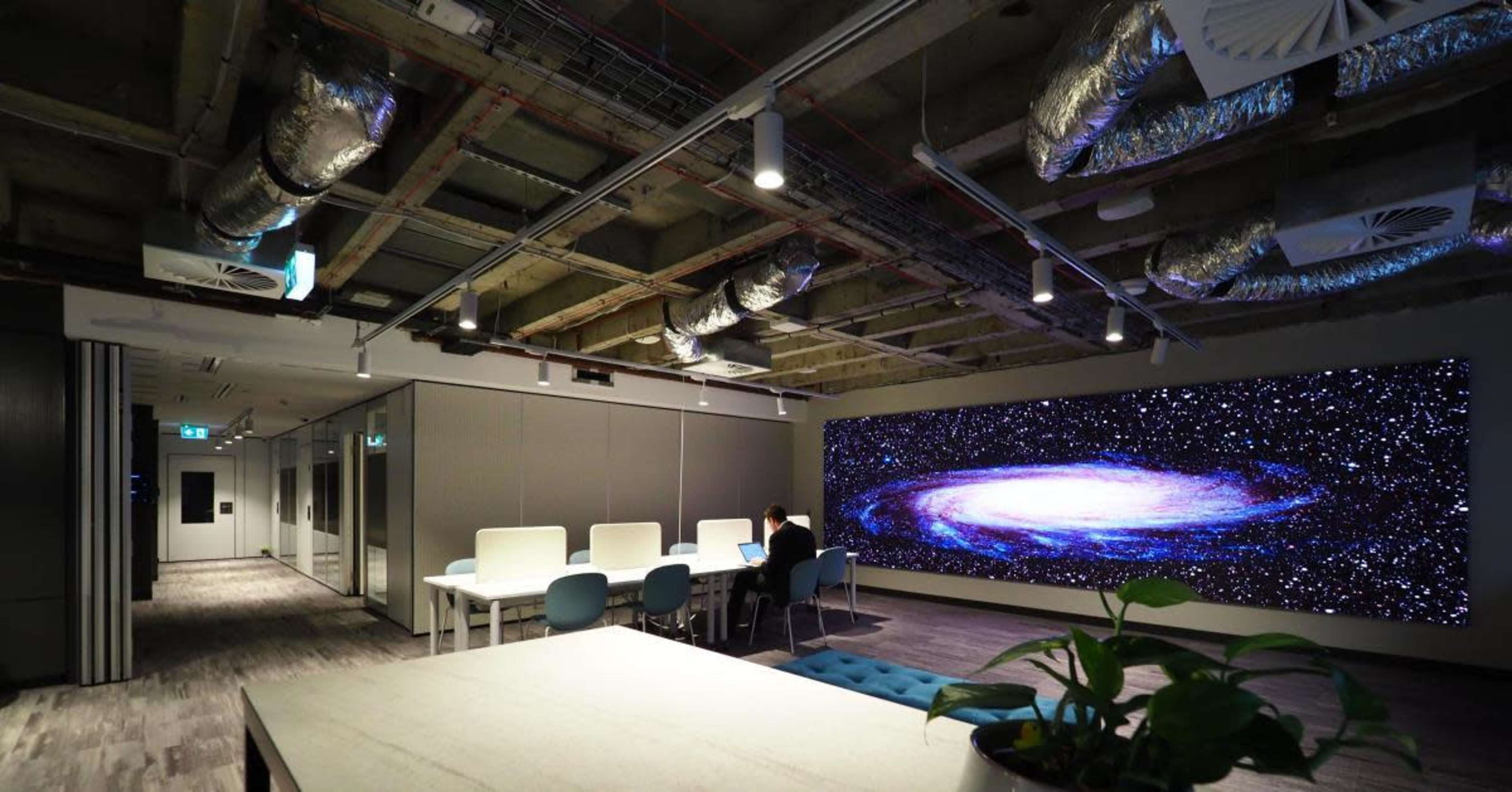 A modern office space with a large screen displaying a galaxy, seating areas, and industrial-style ceiling features.