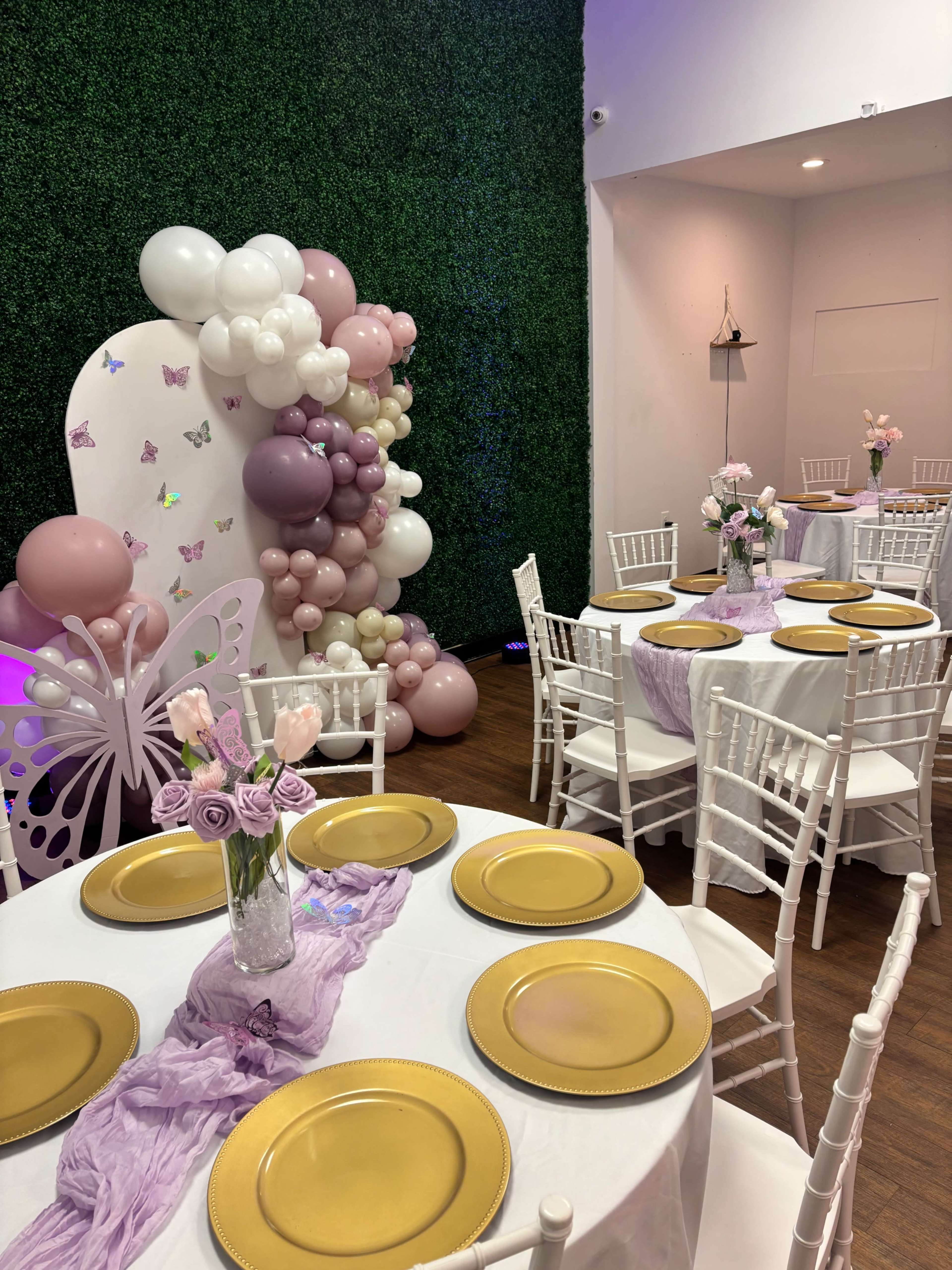 The image shows a decorated event space featuring white tables set with gold plates and lavender table runners, complemented by a backdrop of greenery and a balloon arrangement in shades of pink and white.