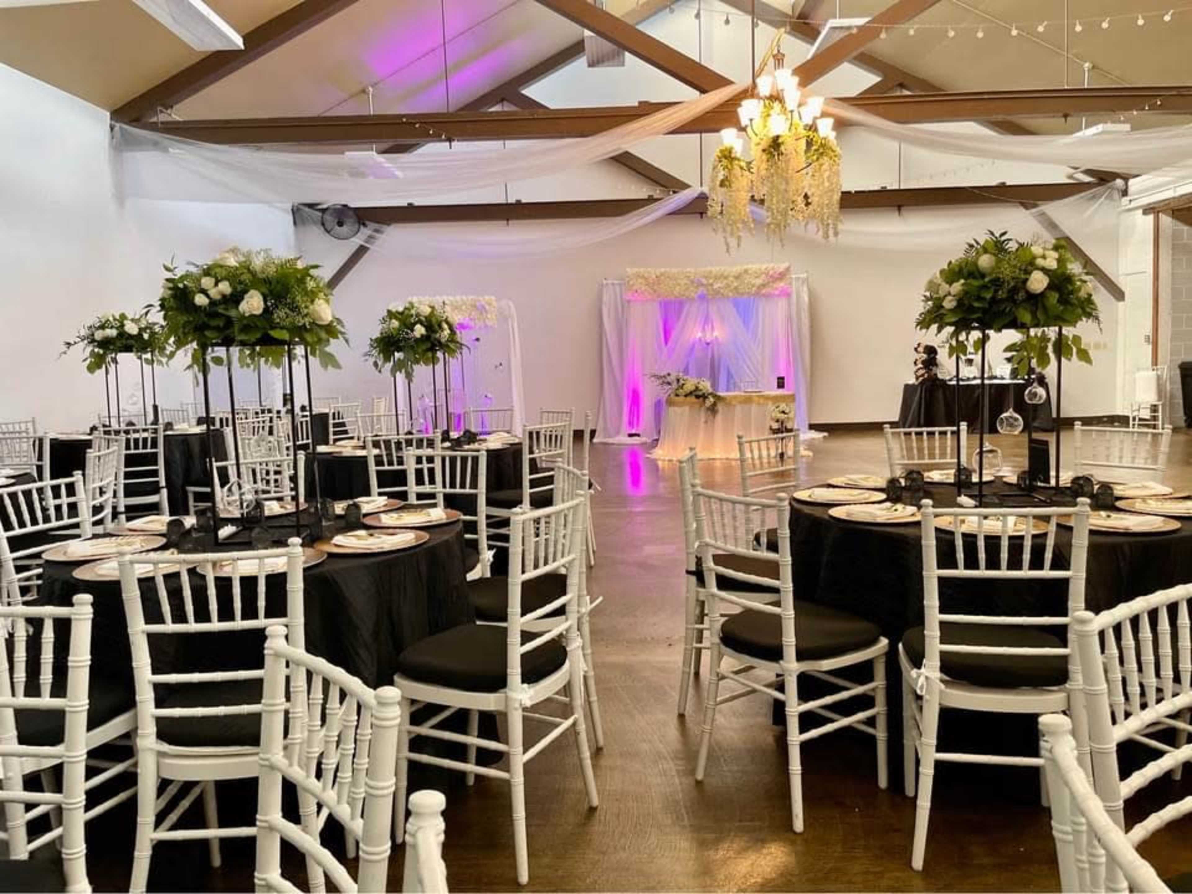 A banquet hall set for an event with round tables adorned with white tableware and tall floral centerpieces, framed by draped fabric and decorative lighting.