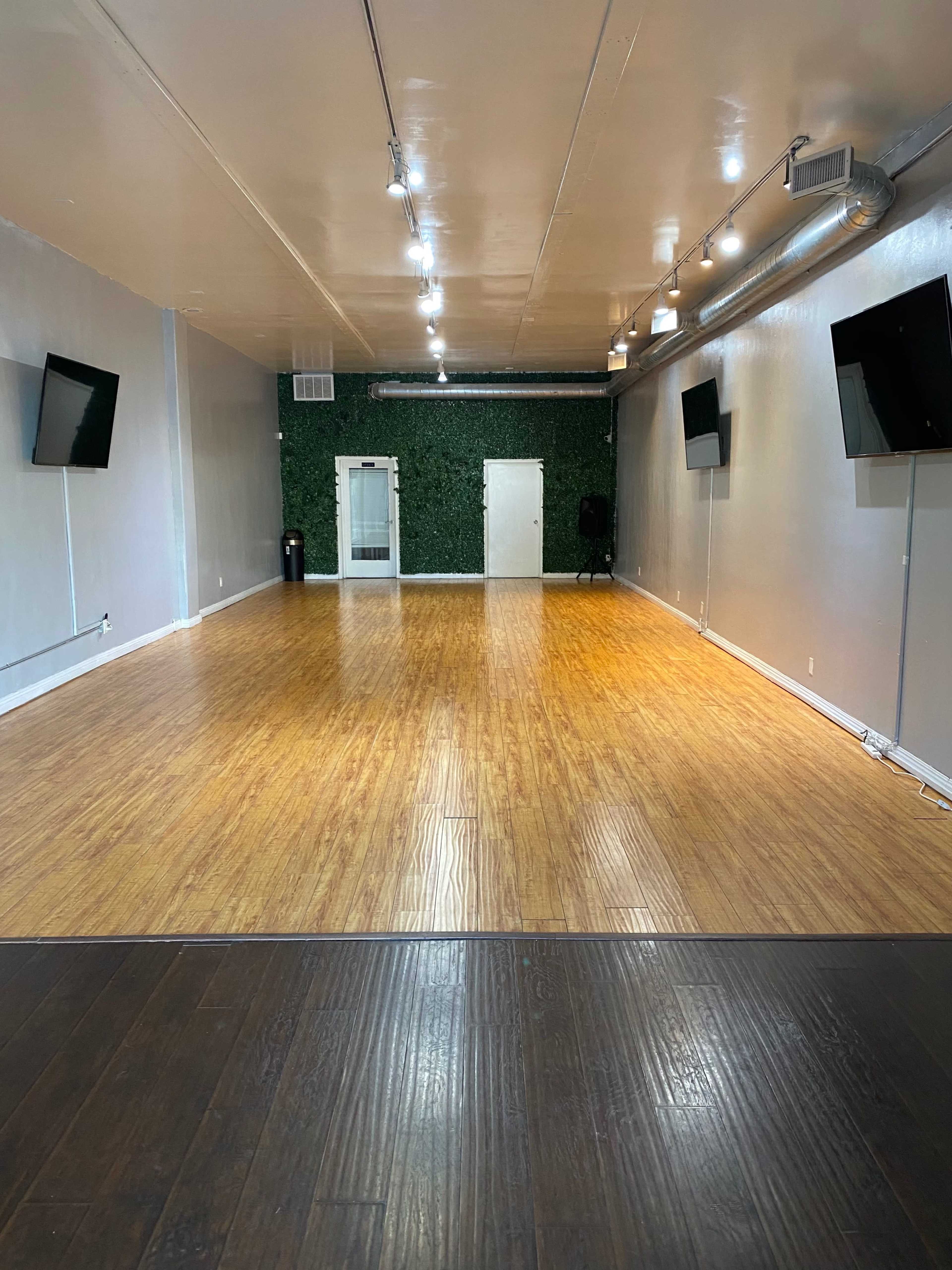 The image shows an empty room with wooden flooring, green wall accents, and two wall-mounted screens.