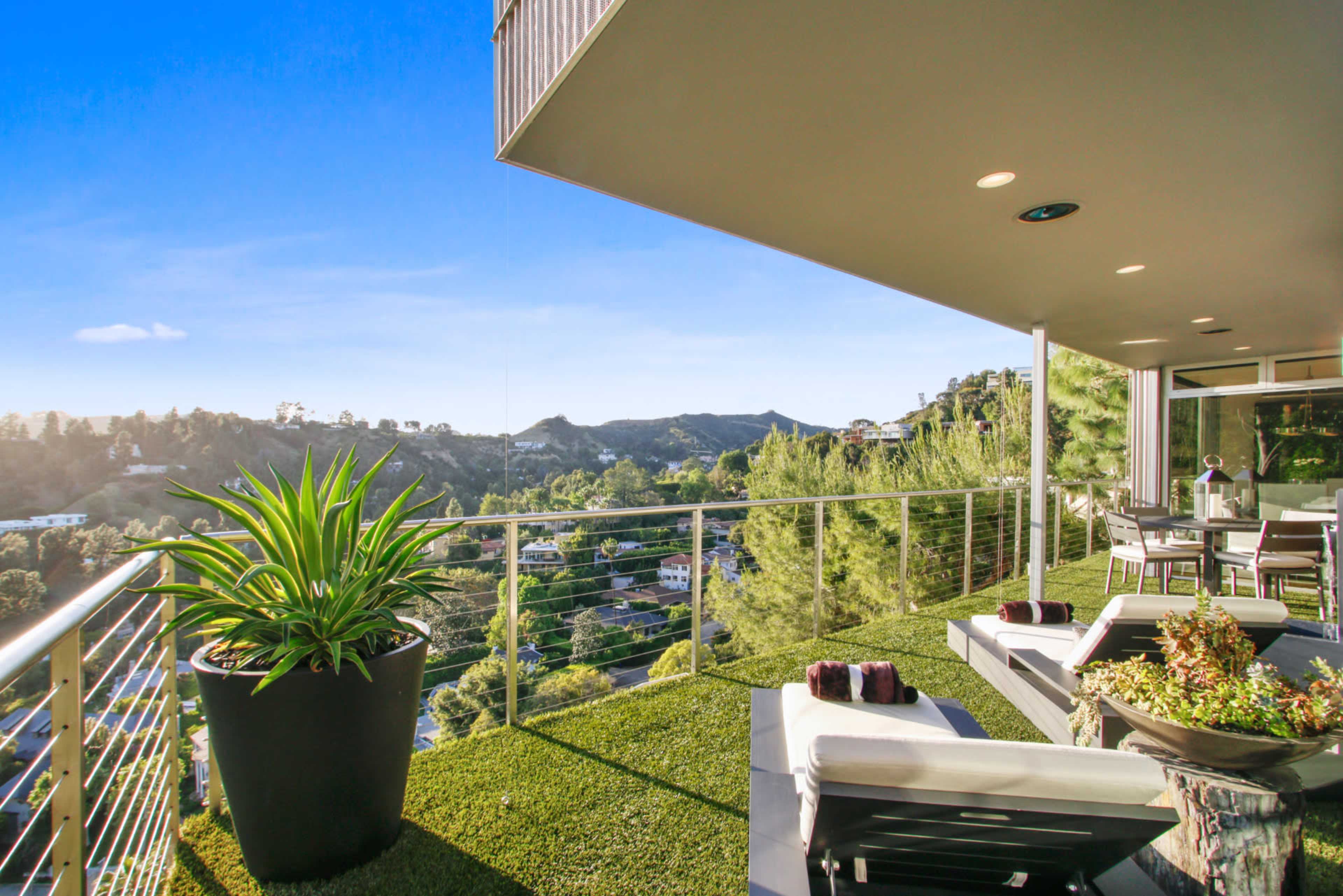 The image shows a modern balcony overlooking a scenic landscape with hills and residential areas, featuring outdoor seating and potted plants.