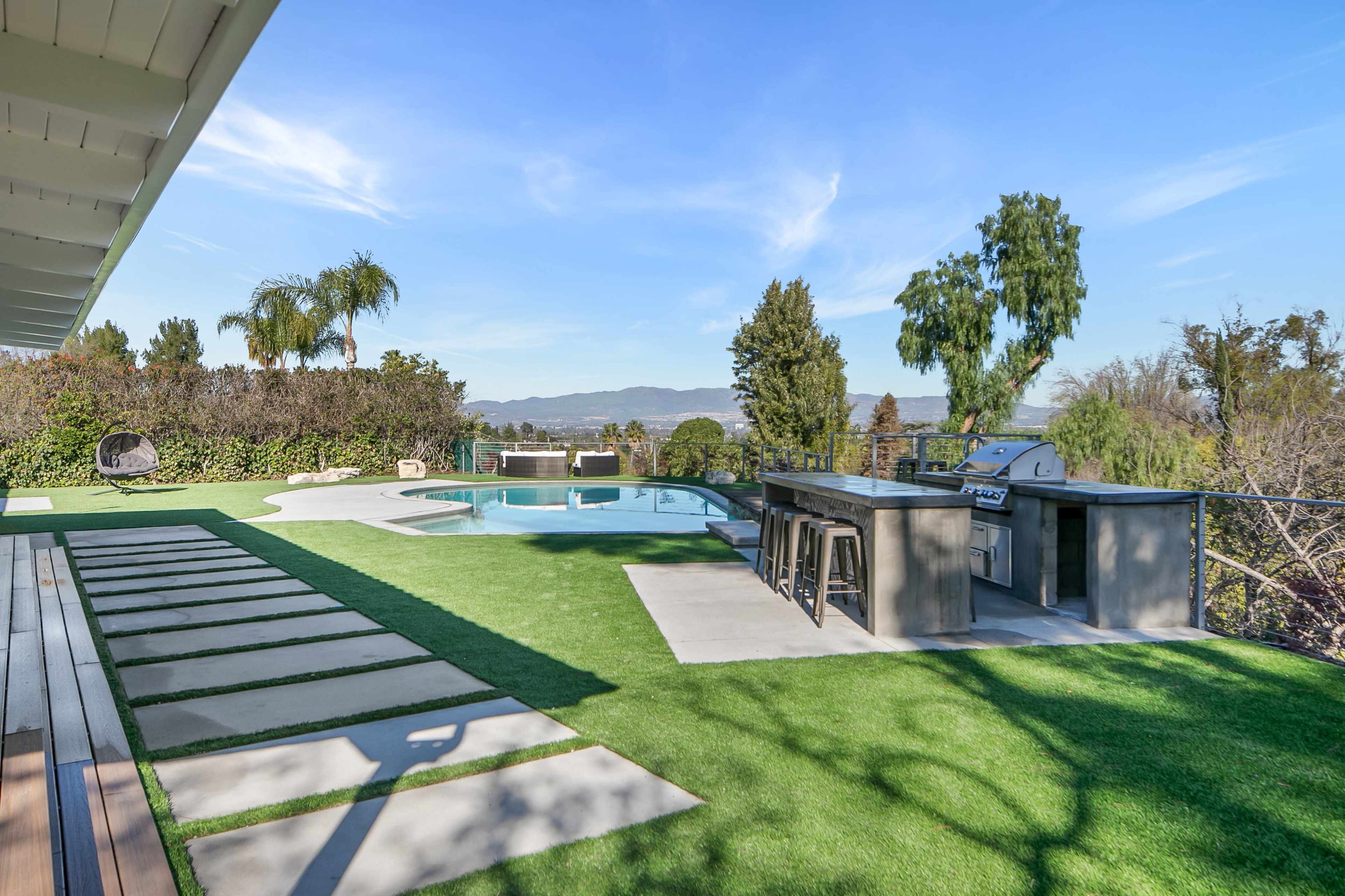 The image shows a backyard with a swimming pool, an outdoor kitchen area, and a landscaped lawn featuring stepping stones.
