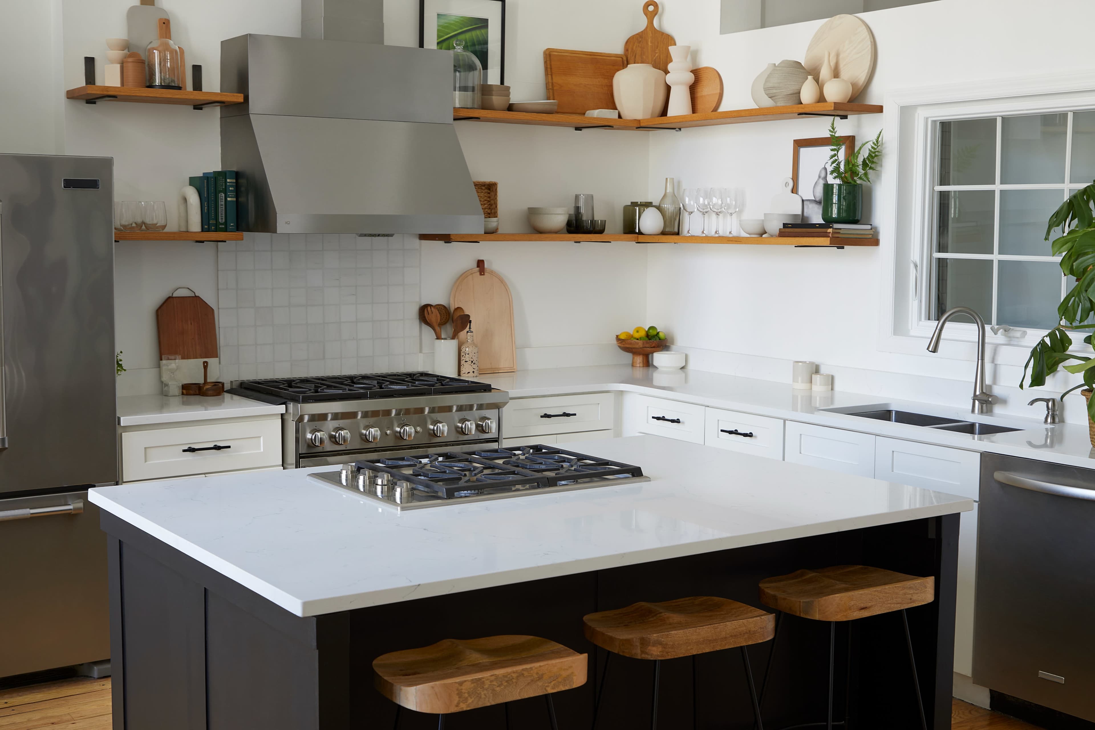 A modern kitchen features a central island with bar stools, stainless steel appliances, and open shelving displaying decorative items and glassware.