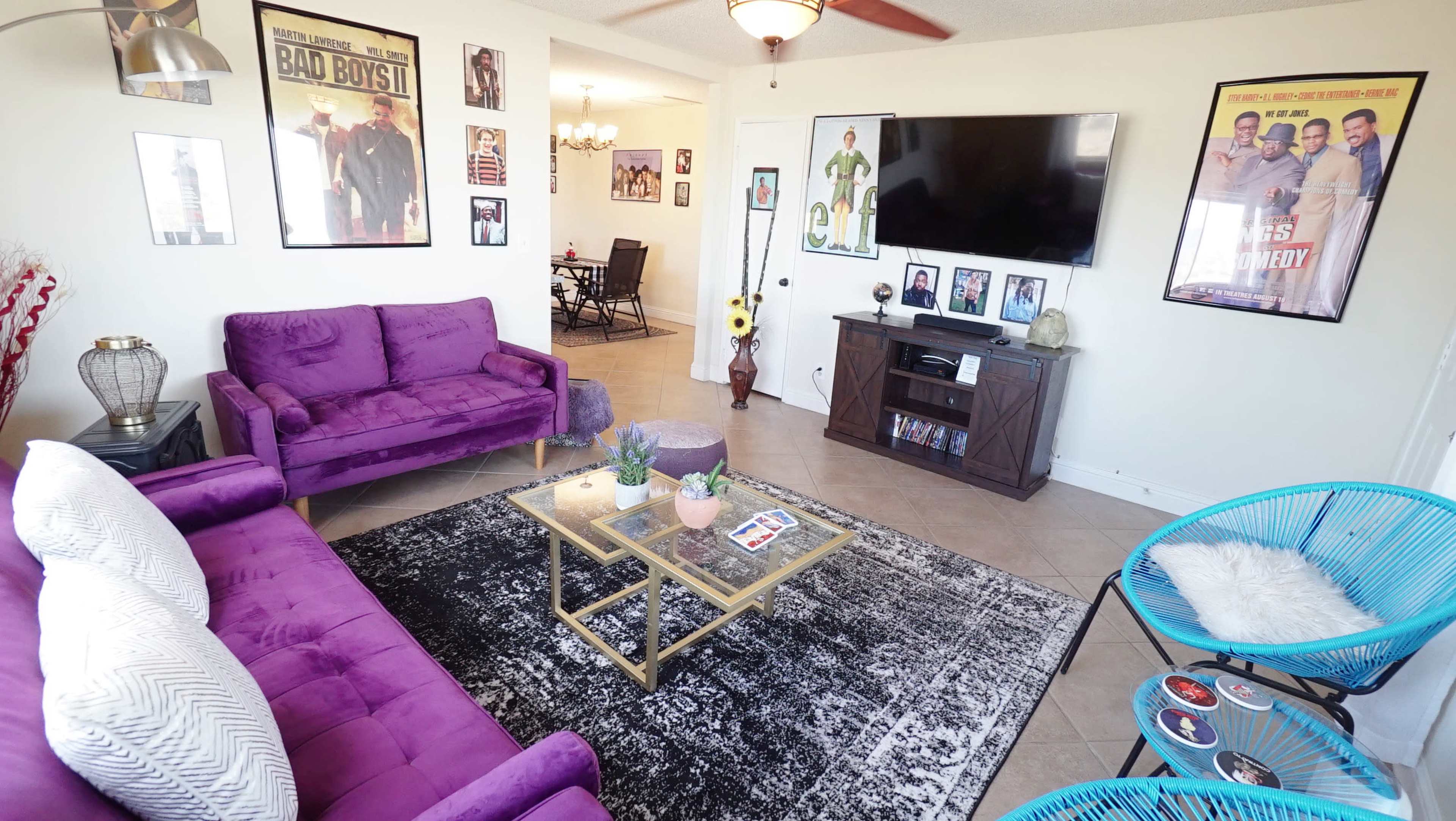 The living room features purple sofas, a coffee table, decorative posters on the walls, and a television mounted above a wooden cabinet.