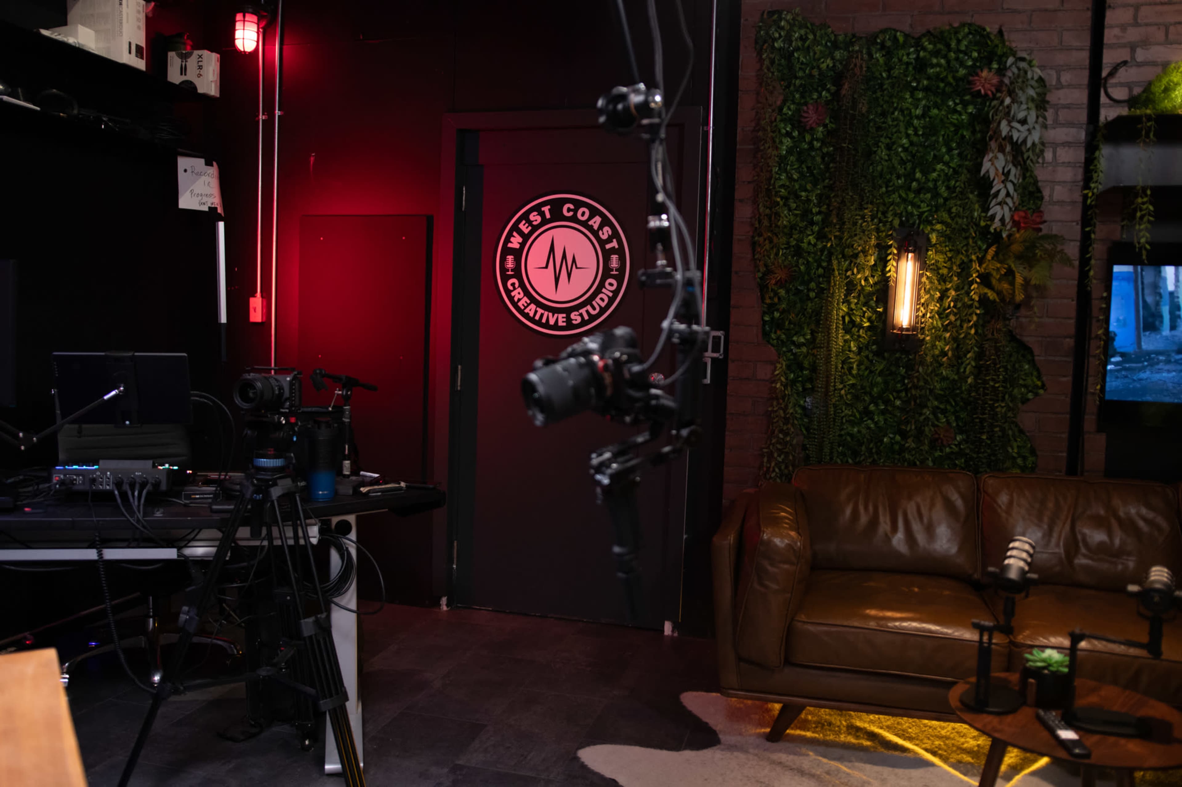 A dimly lit studio with a camera setup, a brown leather couch, and a door labeled "West Coast Creative Studio" adorned with a logo.