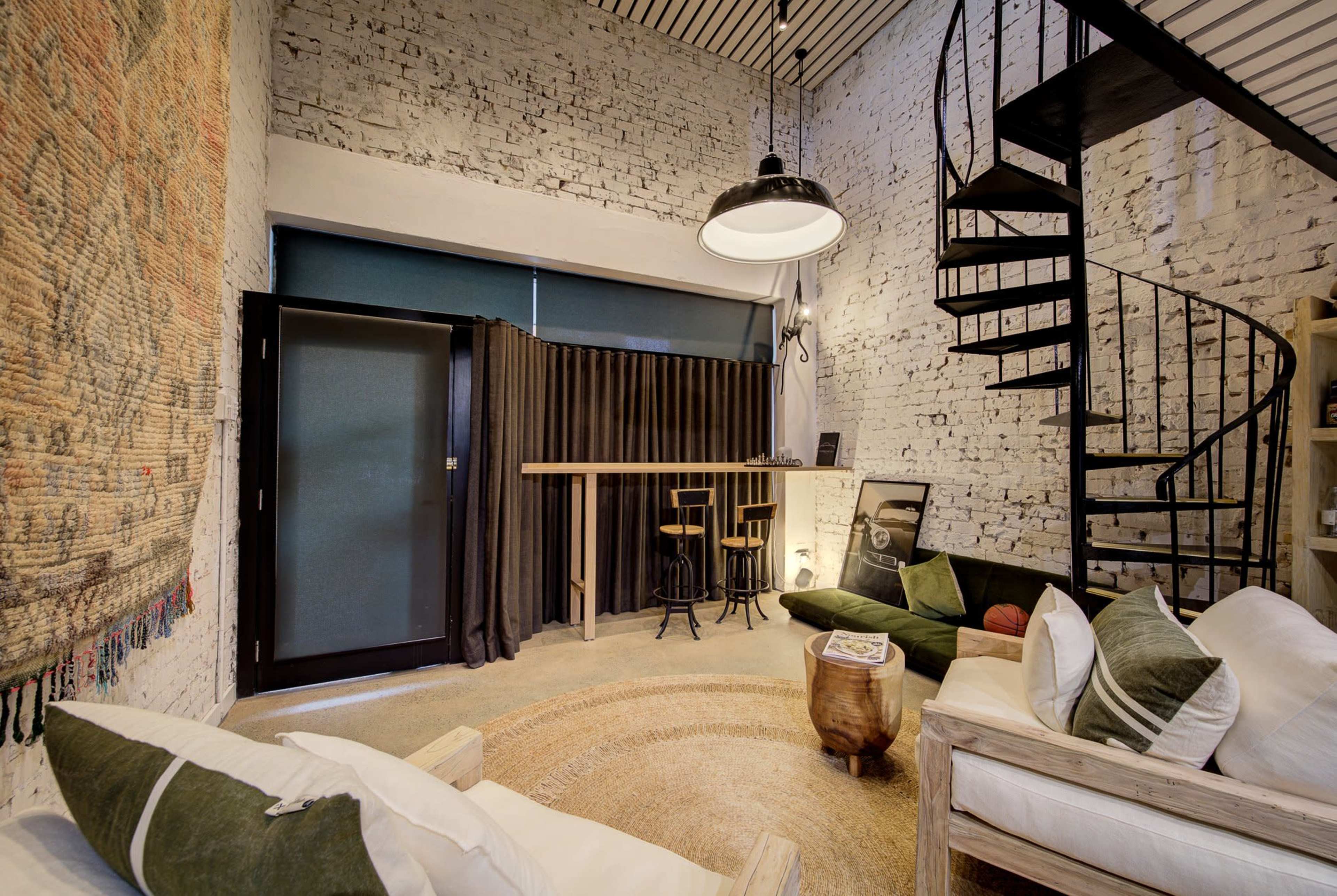 The image shows a modern living space with exposed brick walls, a spiral staircase, a small bar area with two stools, and a circular rug in the center.