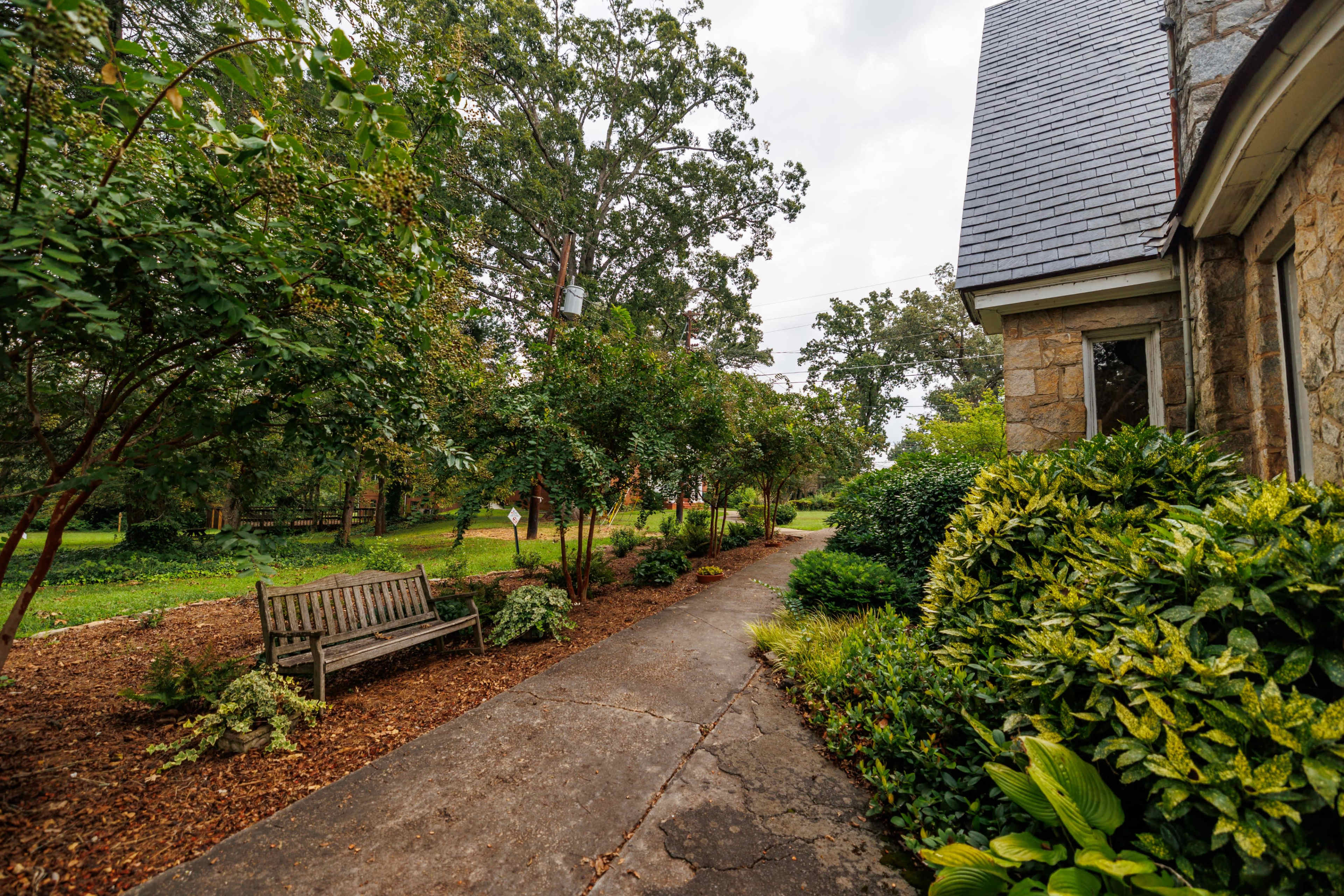 The image shows a landscaped pathway lined with shrubs and trees, leading up to a stone building.