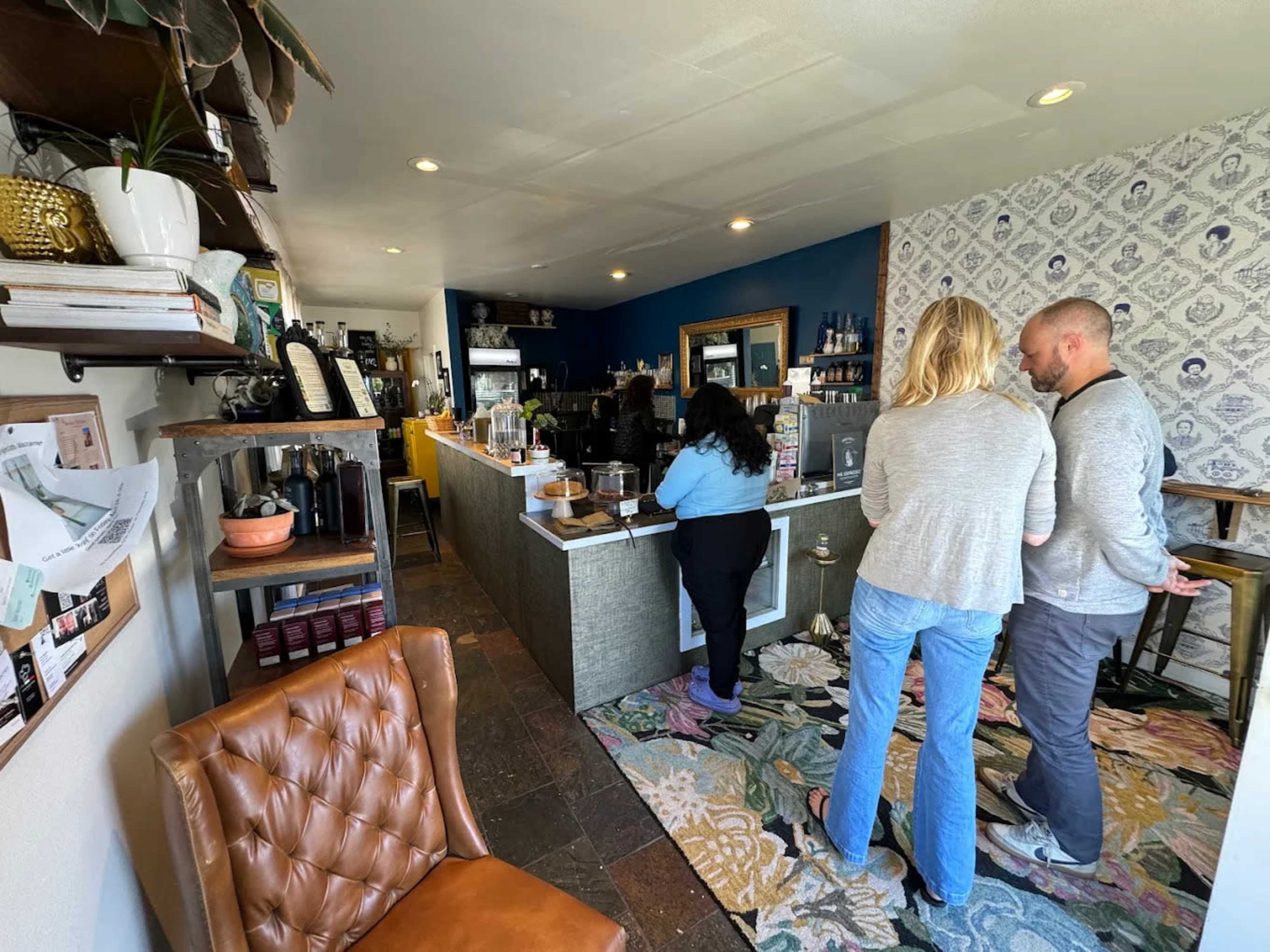 A coffee shop interior features a customer at the counter with two people standing nearby, surrounded by decor and displayed merchandise.