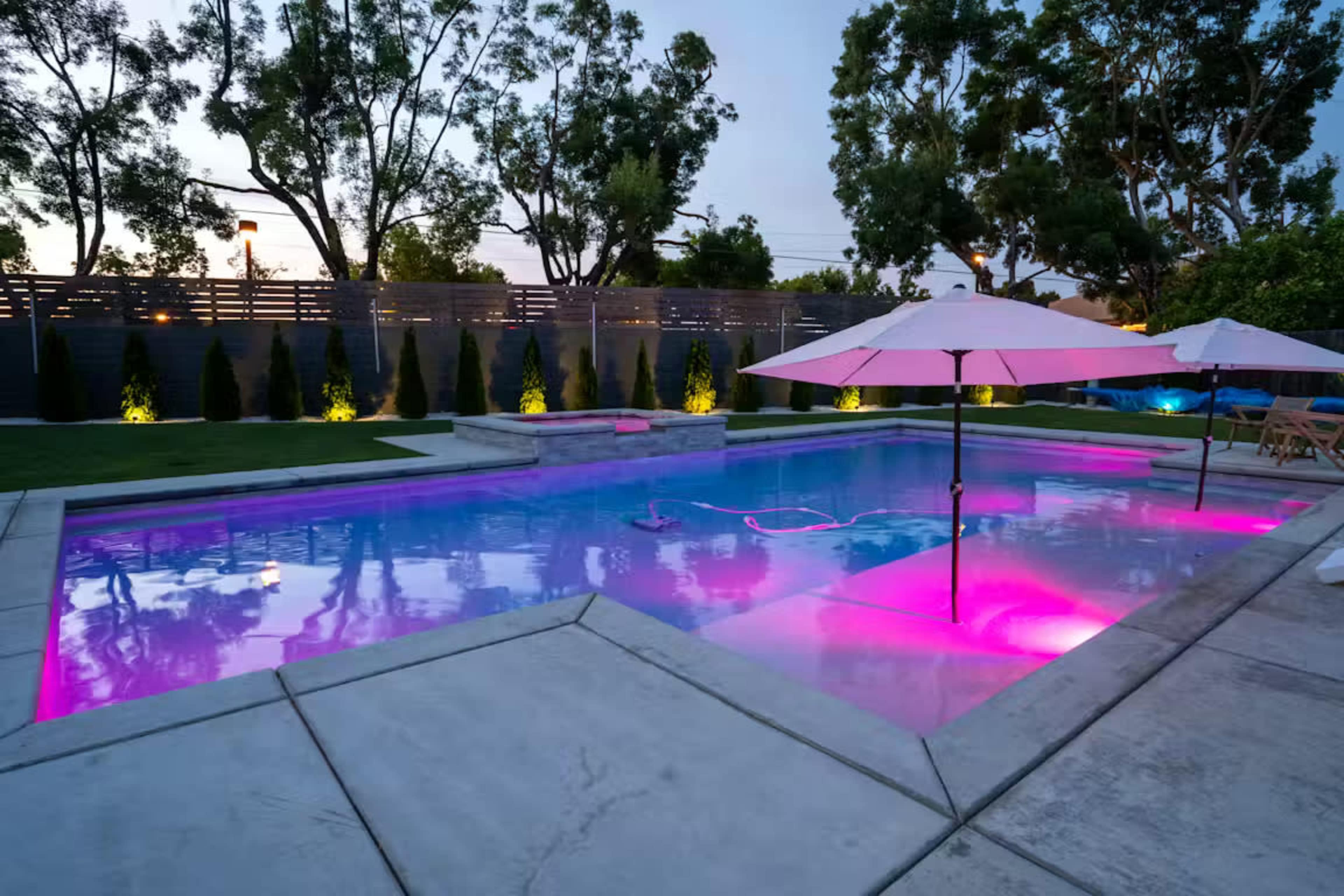 The image shows a modern swimming pool illuminated with pink lights, surrounded by trees and landscaped greenery at dusk.