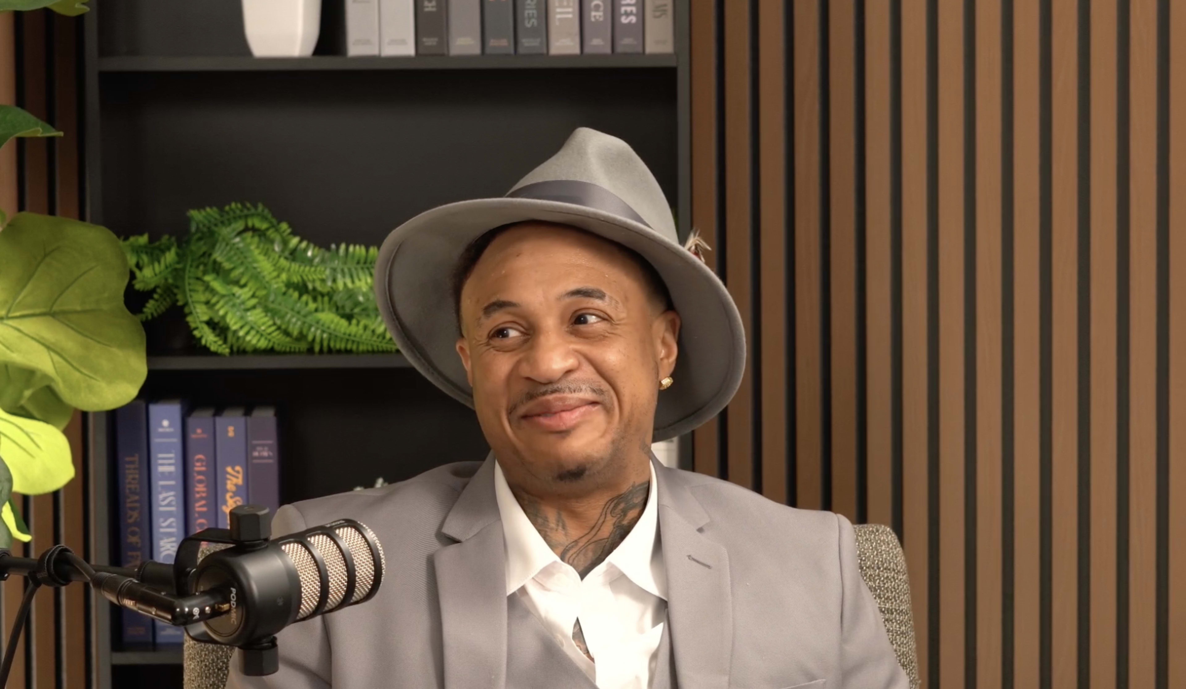 A man wearing a gray suit and hat smiles while sitting in front of a microphone and a bookshelf.