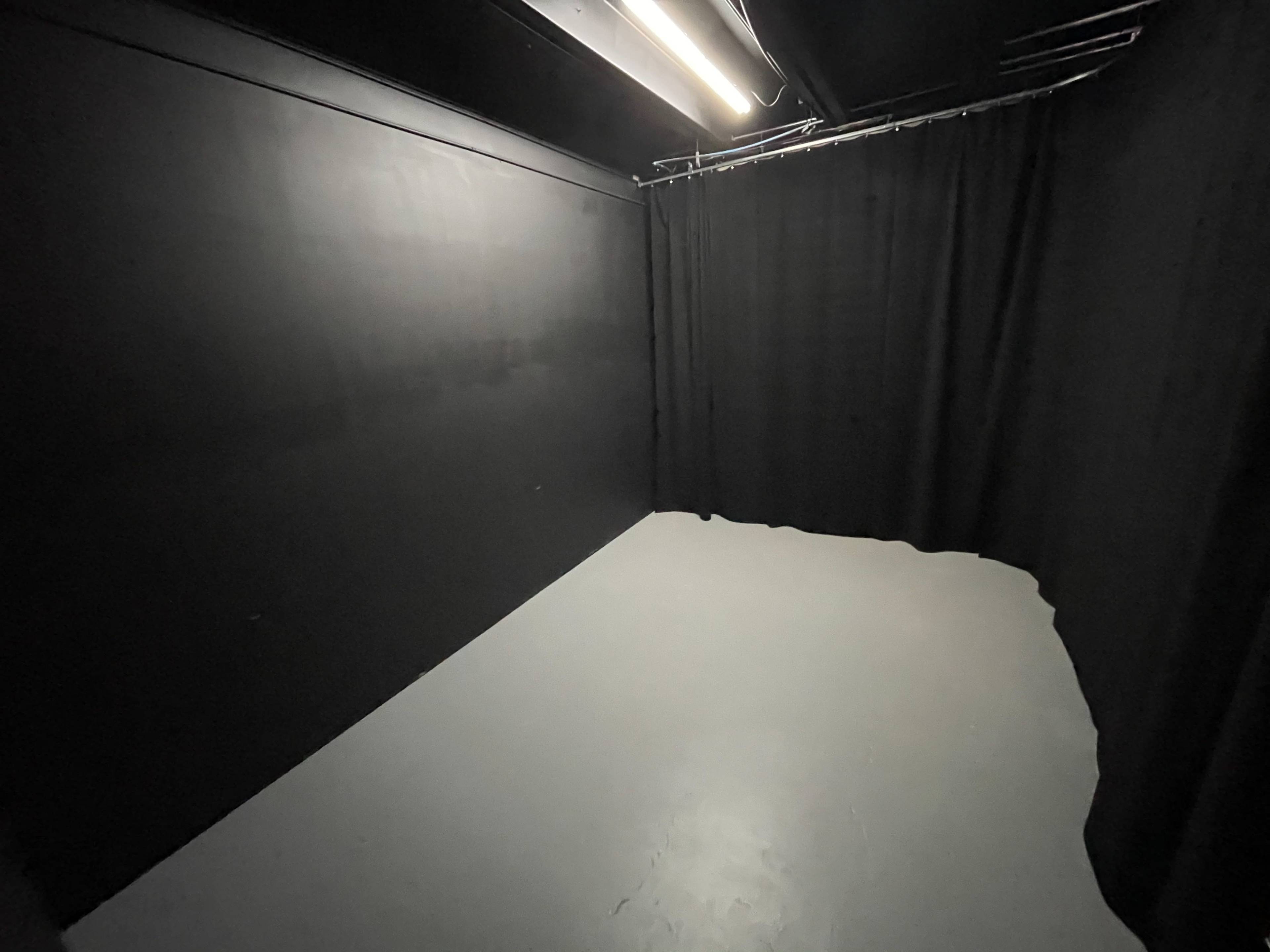 The image shows a dimly lit room with black walls and a gray floor, featuring a curtain along one side.