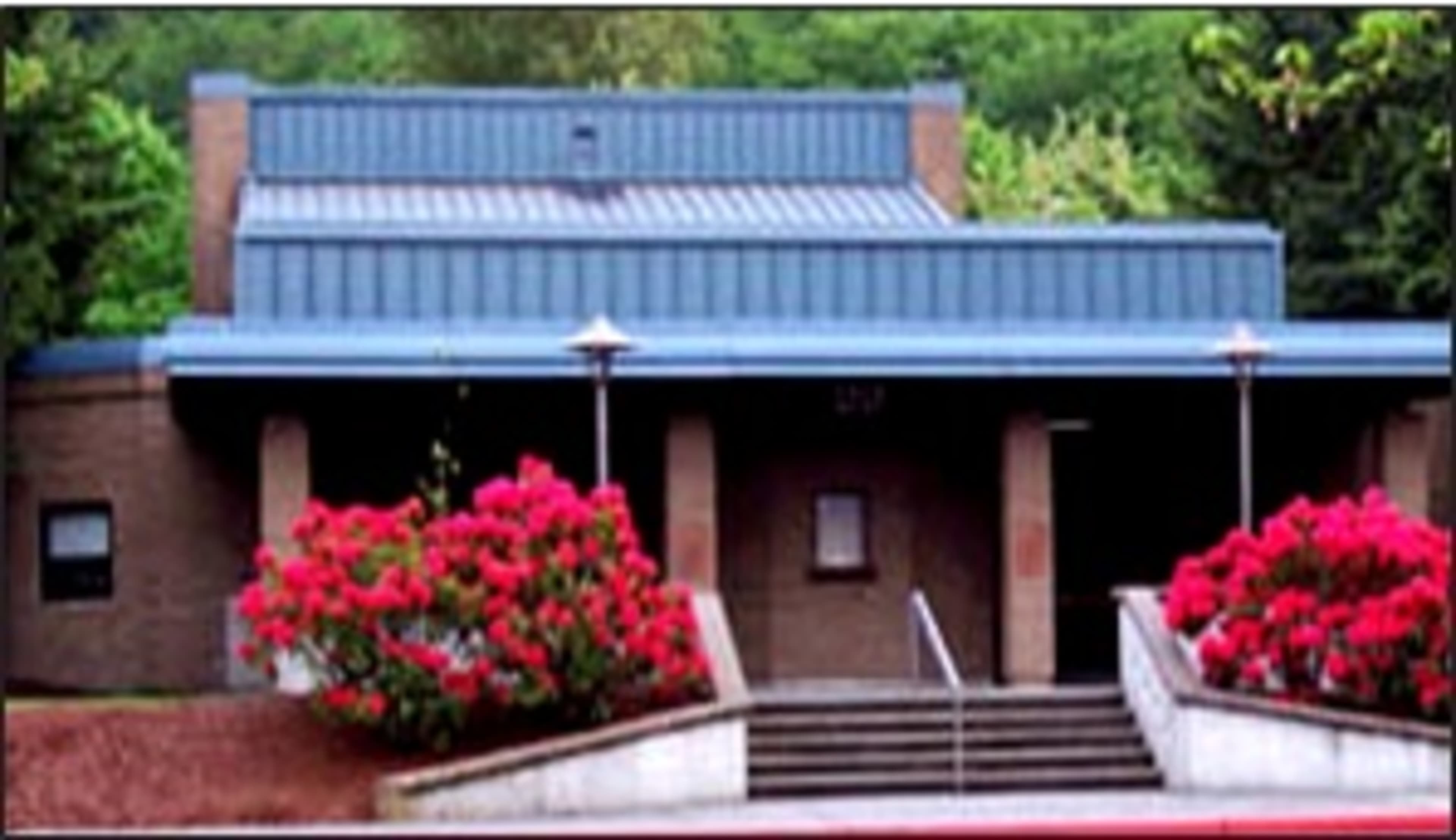 A building with a blue metal roof and pink flowering shrubs in front, featuring a set of stairs leading to the entrance.