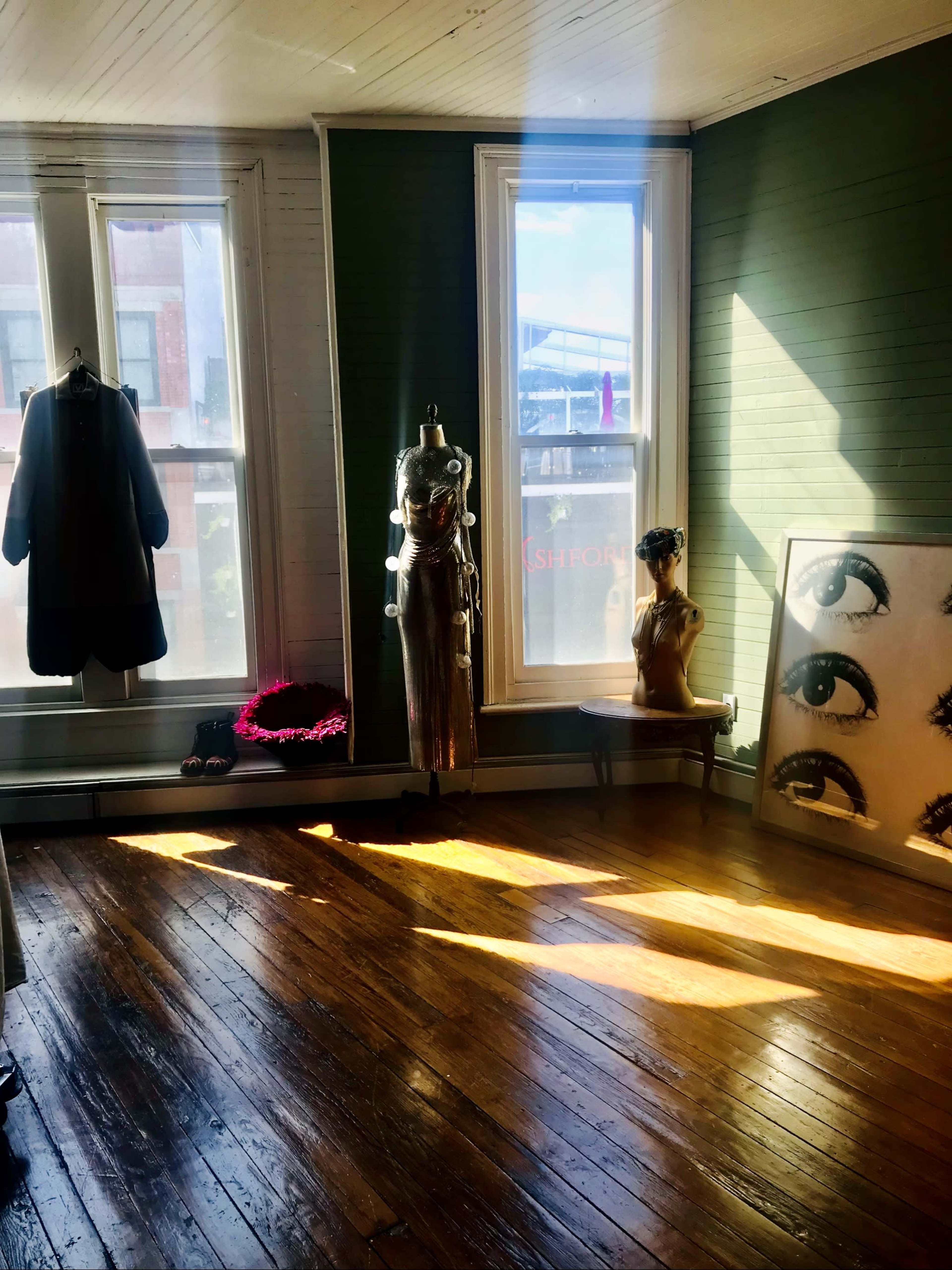 The room features two large windows, a mannequin dressed in gold fabric, a decorative head with a vision-related theme, and a coat hanging on a rack.