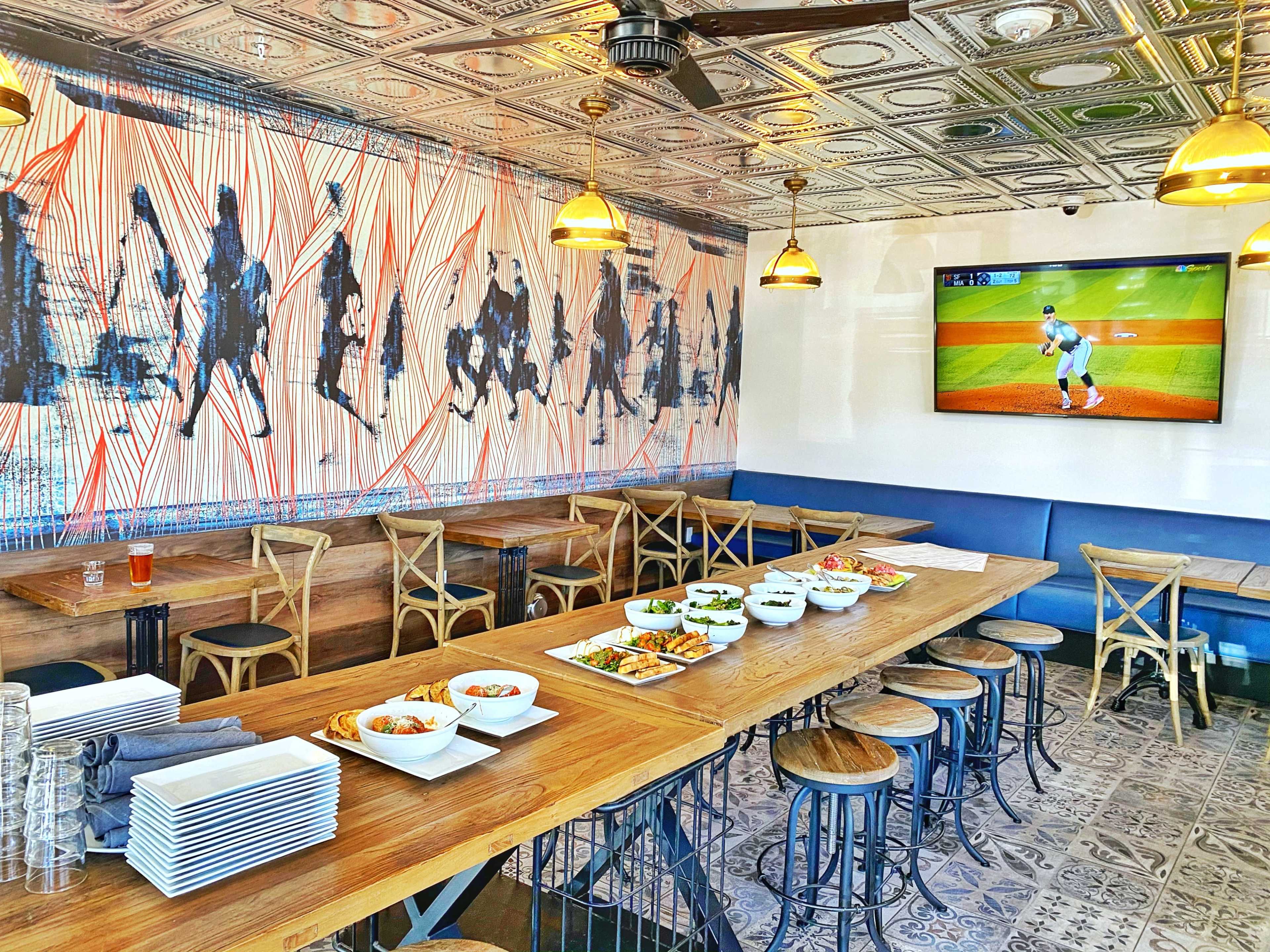 A spacious restaurant interior features a long wooden table set with various dishes, surrounded by wooden chairs and a wall mural depicting figures, with a television displaying a sports event.