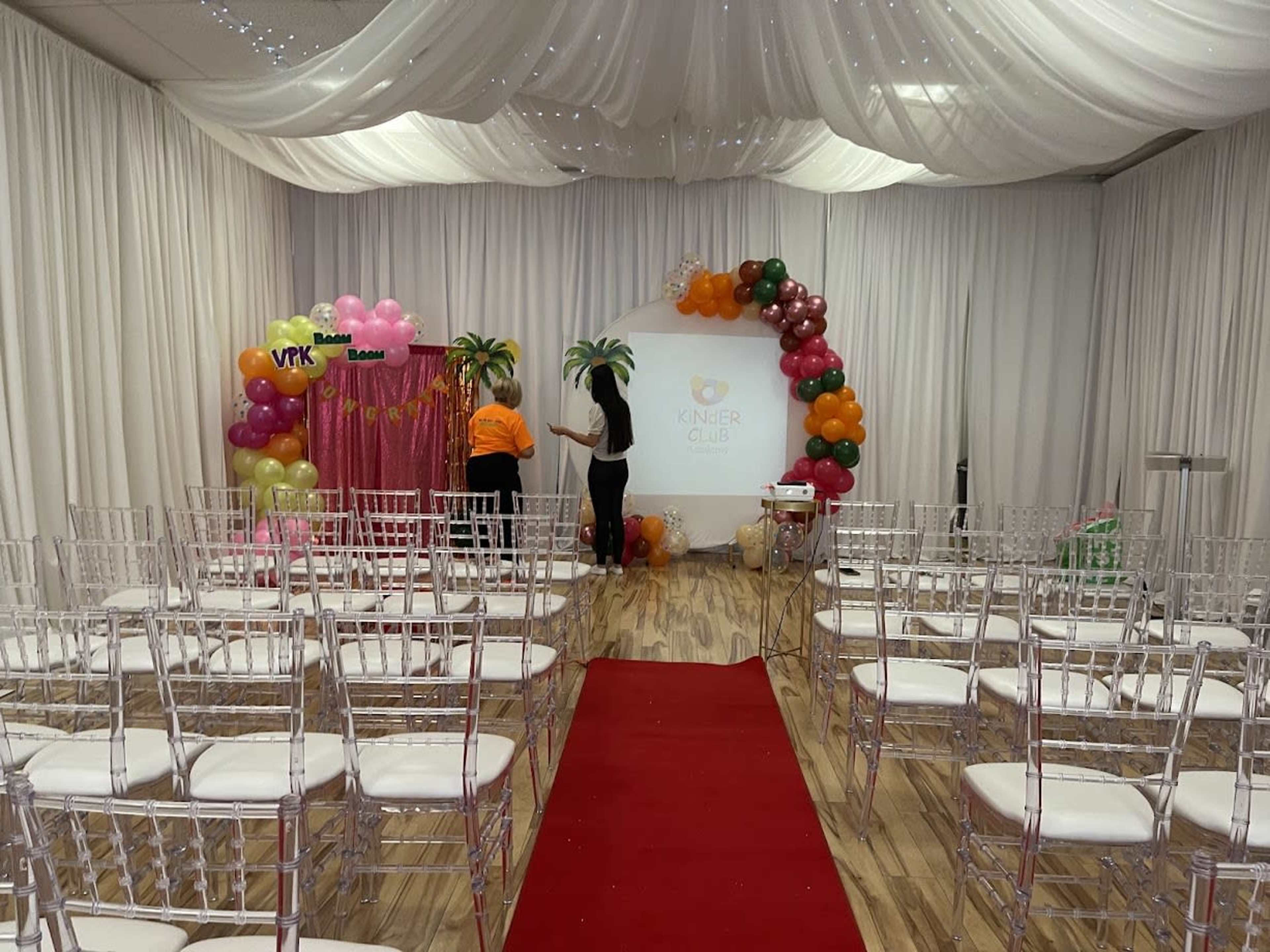 The image shows an event setup with a red carpet leading to a decorated stage featuring balloons and a backdrop, surrounded by rows of clear chairs.