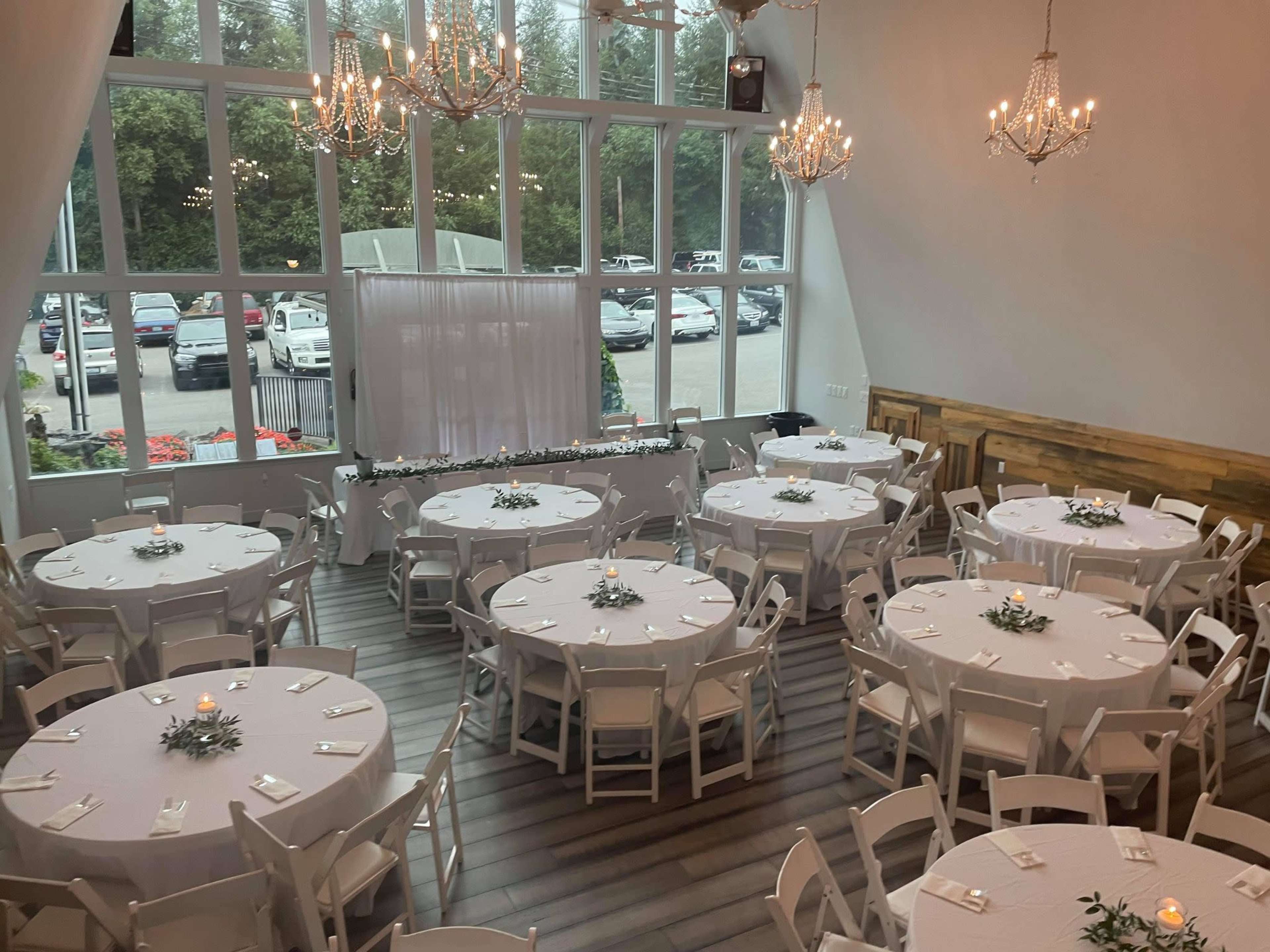 A banquet hall with several round tables set for an event, featuring white tablecloths and floral centerpieces, under chandeliers and large windows.