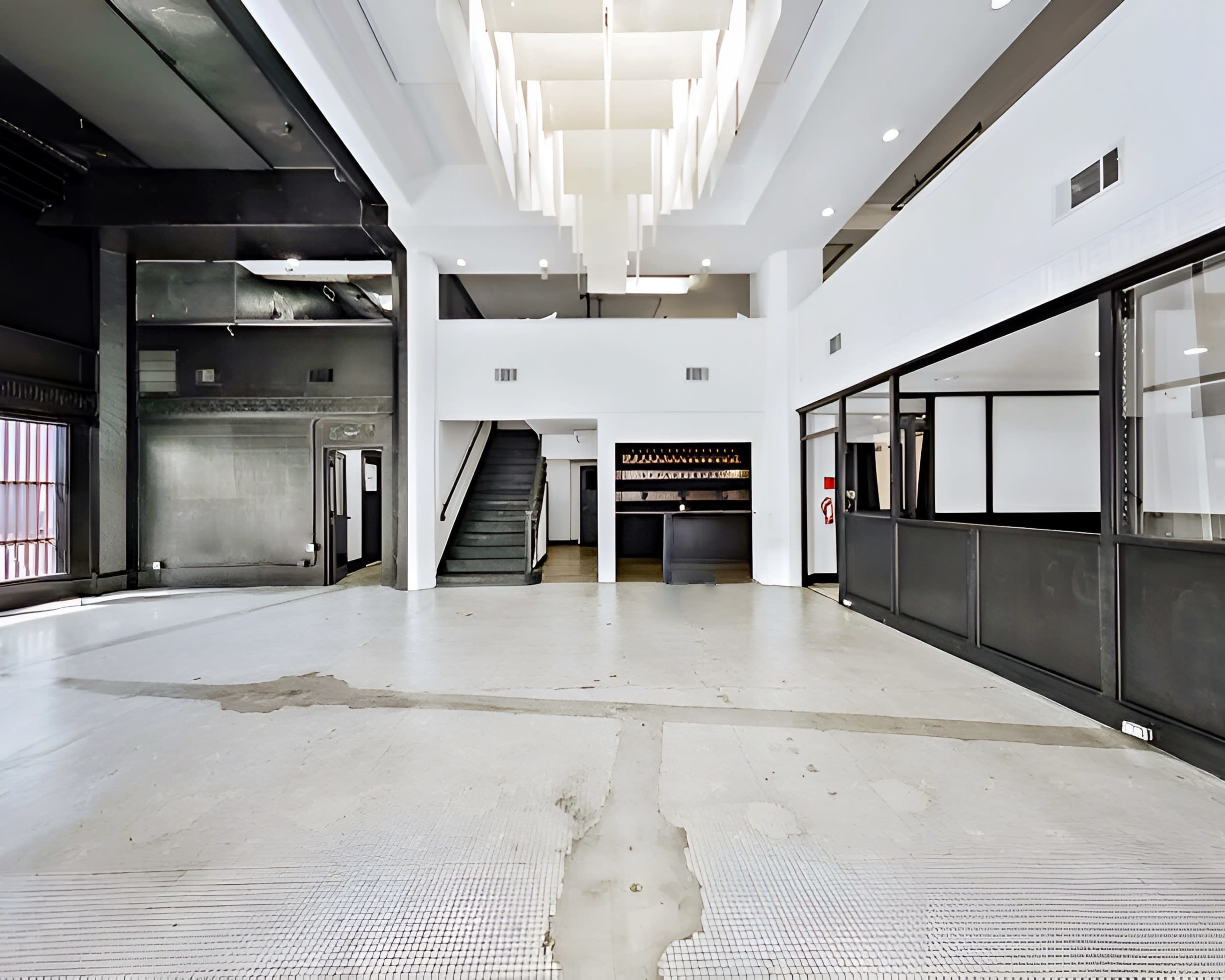 The image shows an empty commercial space with a large, open floor area, a staircase in the background, and a modern lighting fixture.