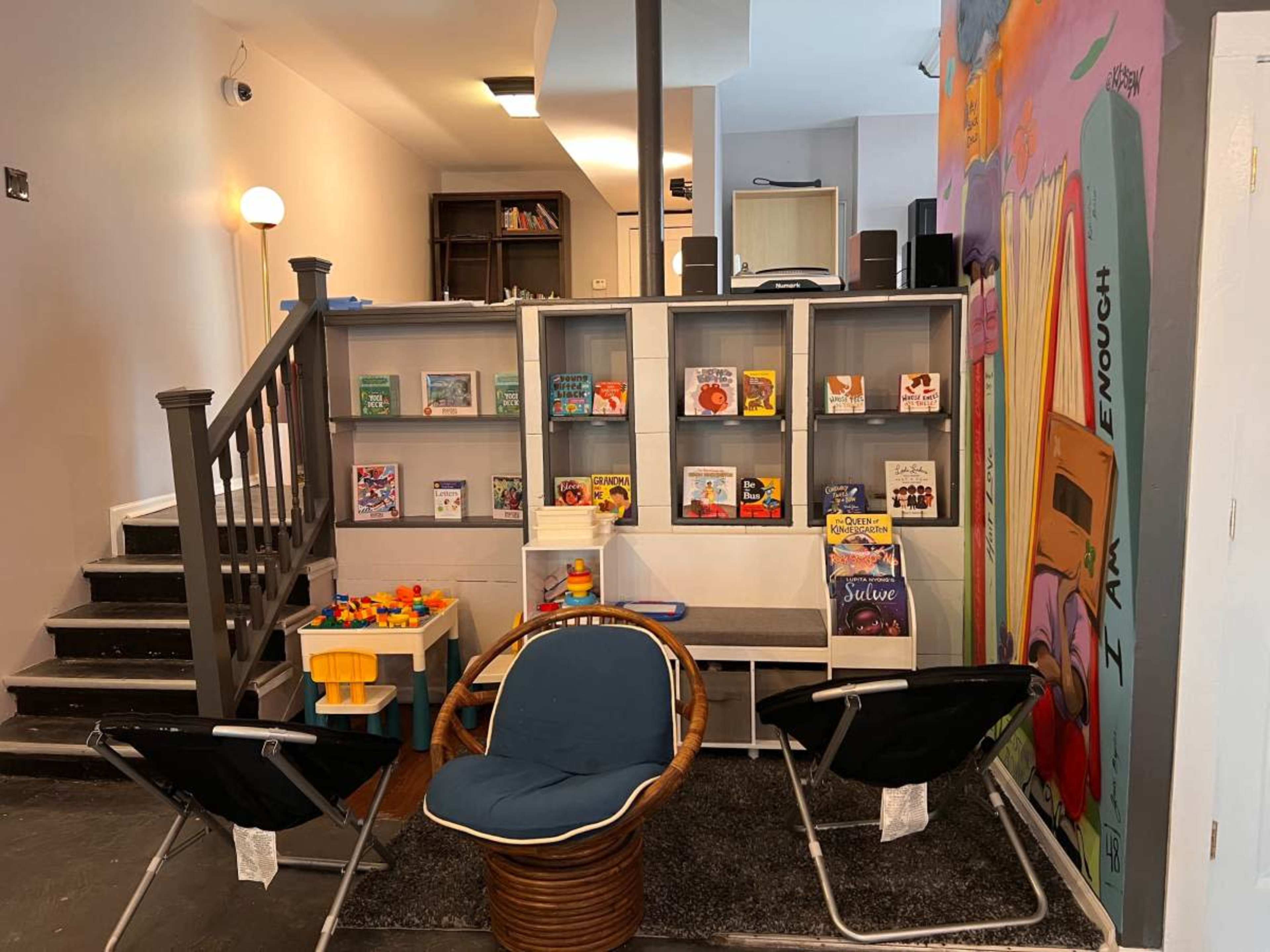 The image shows a cozy reading nook with bookshelves filled with various books and a small seating area featuring two chairs and colorful children's toys.