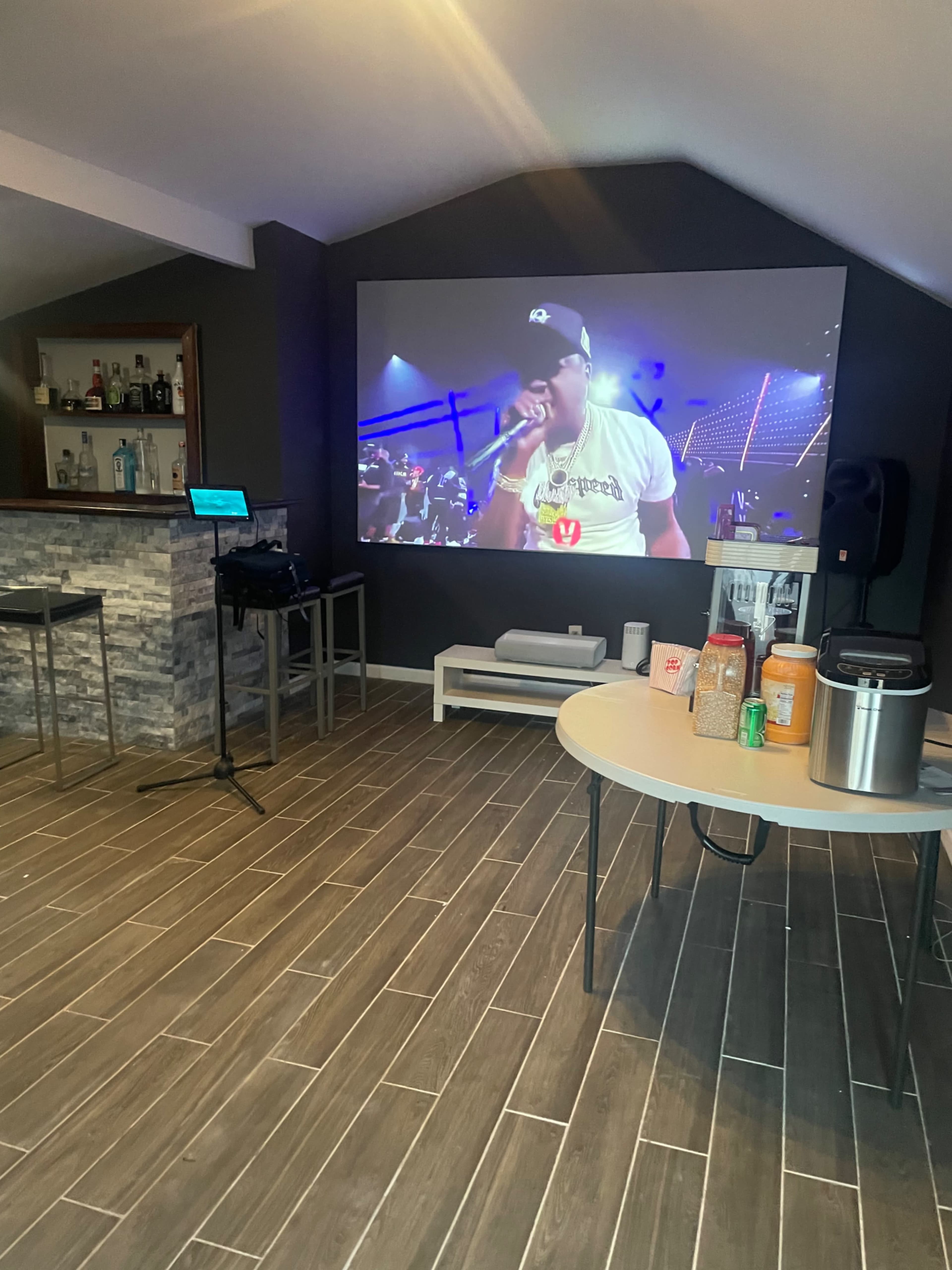 A home entertainment area with a large screen displaying a performer, a bar counter with various drinks, and a table with snacks and a beverage dispenser.