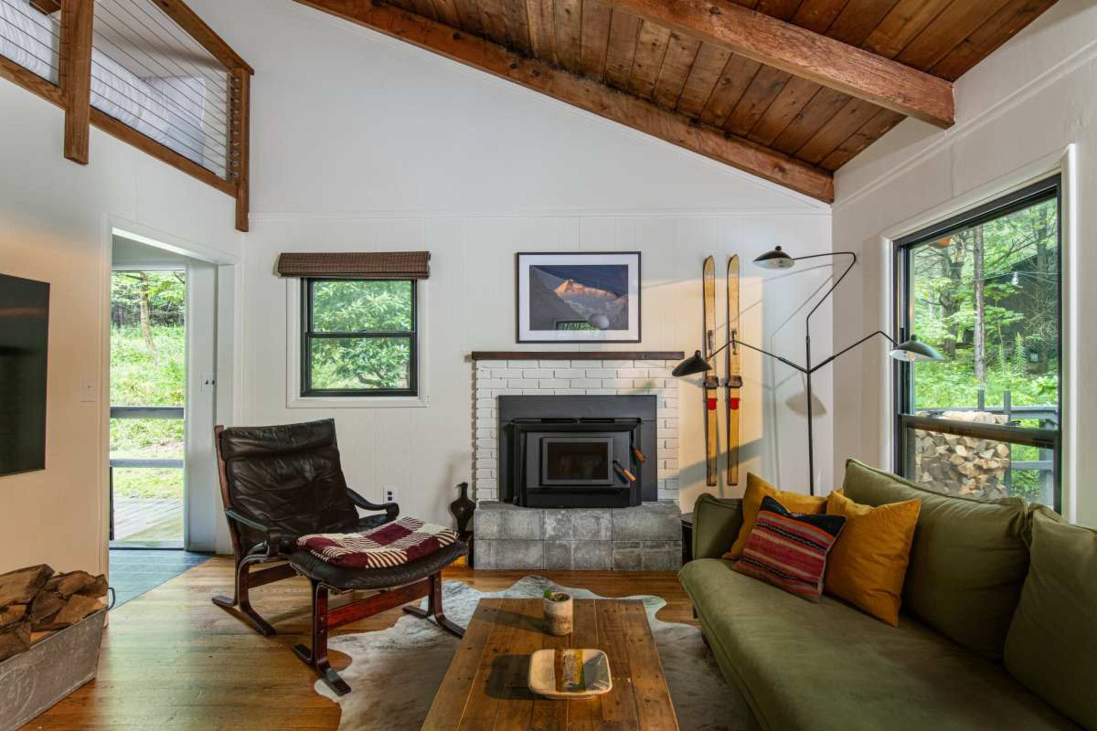 The image shows a cozy living room featuring a fireplace, wooden beams on the ceiling, a green sofa, and ski equipment mounted on the wall.