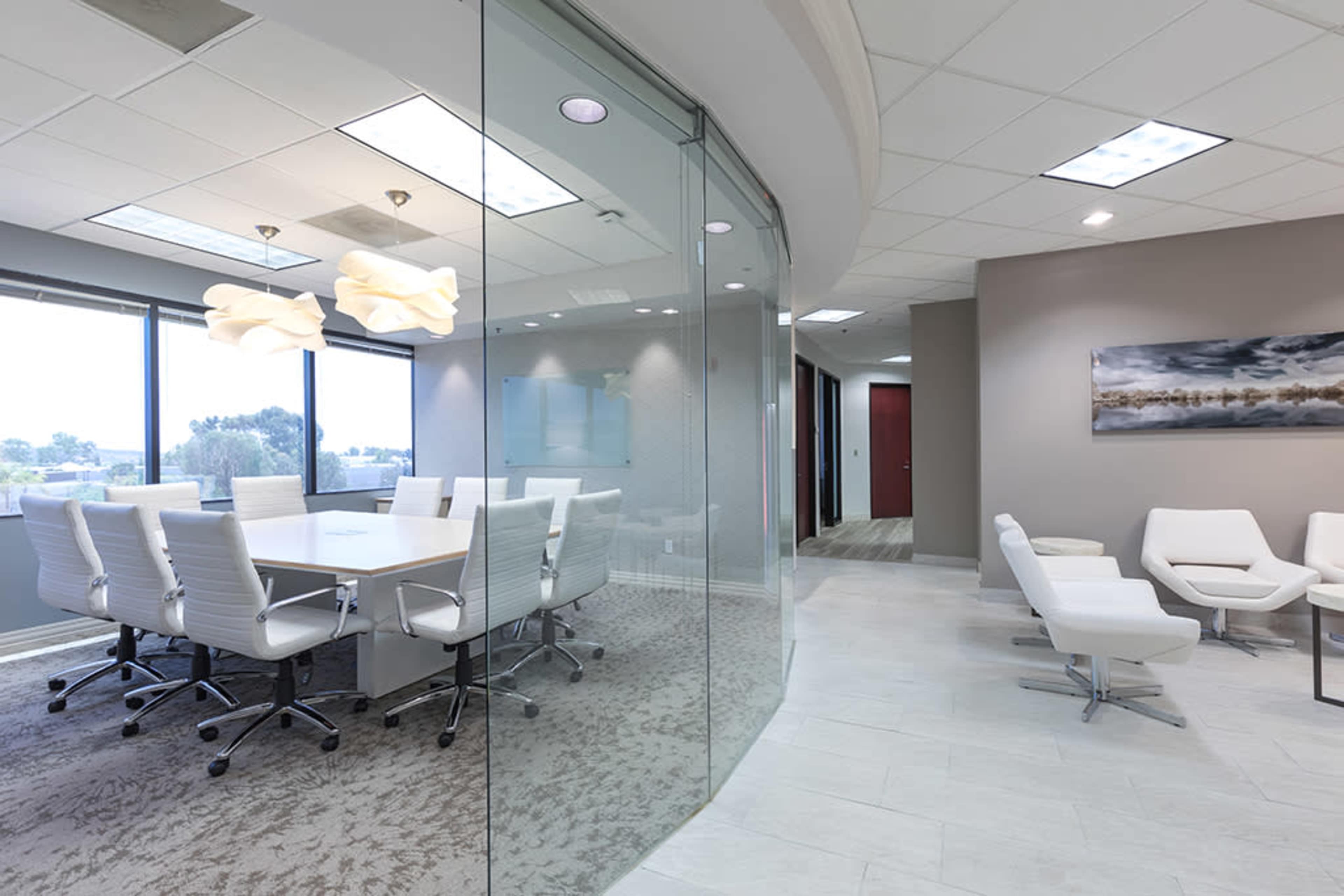 A modern conference room with a large table surrounded by white chairs, overlooking a city view through a glass wall.