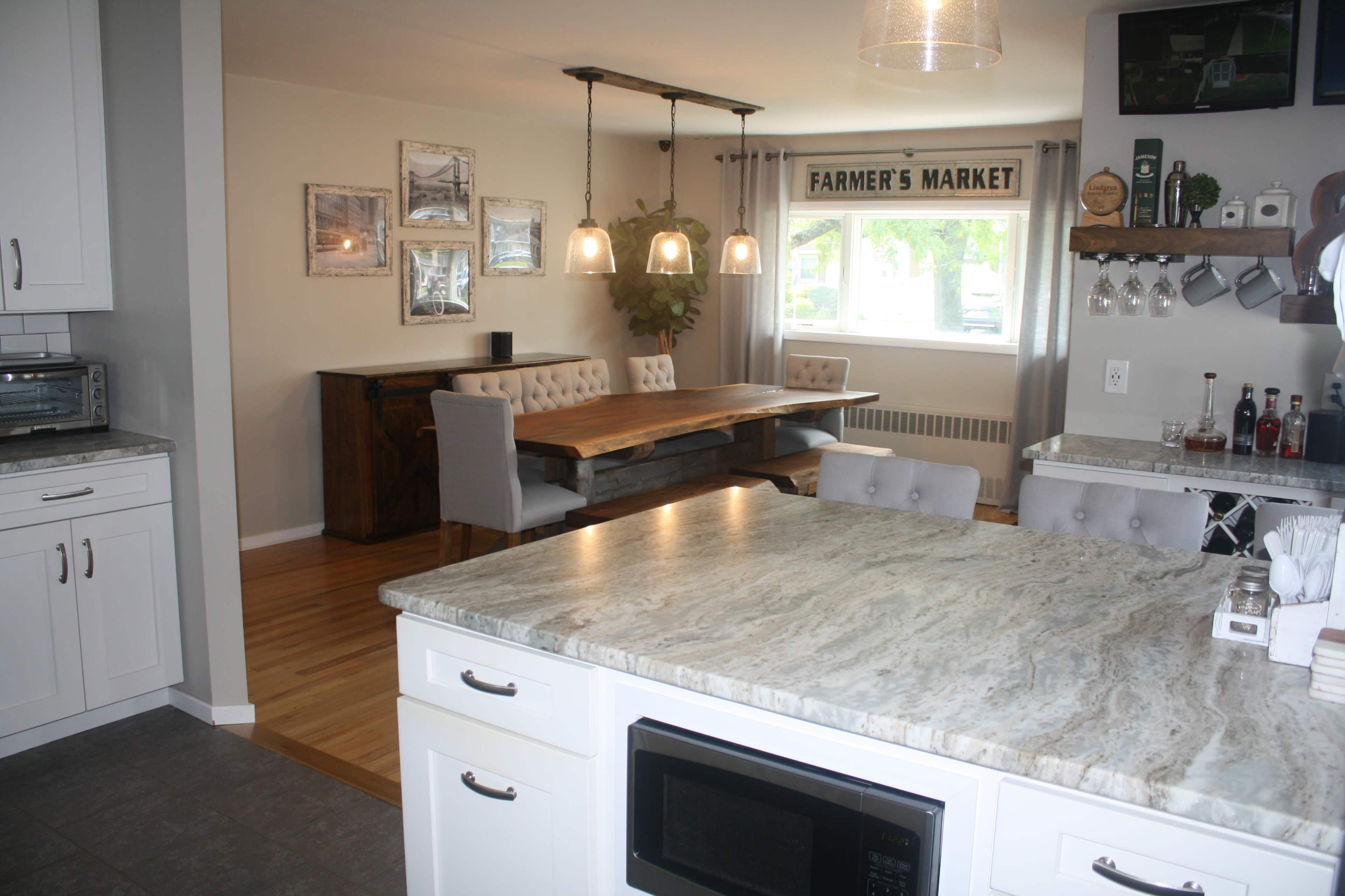 A kitchen with a marble countertop and white cabinets, overlooking a dining area featuring a wooden table and grey chairs under a sign reading "FARMER'S MARKET."