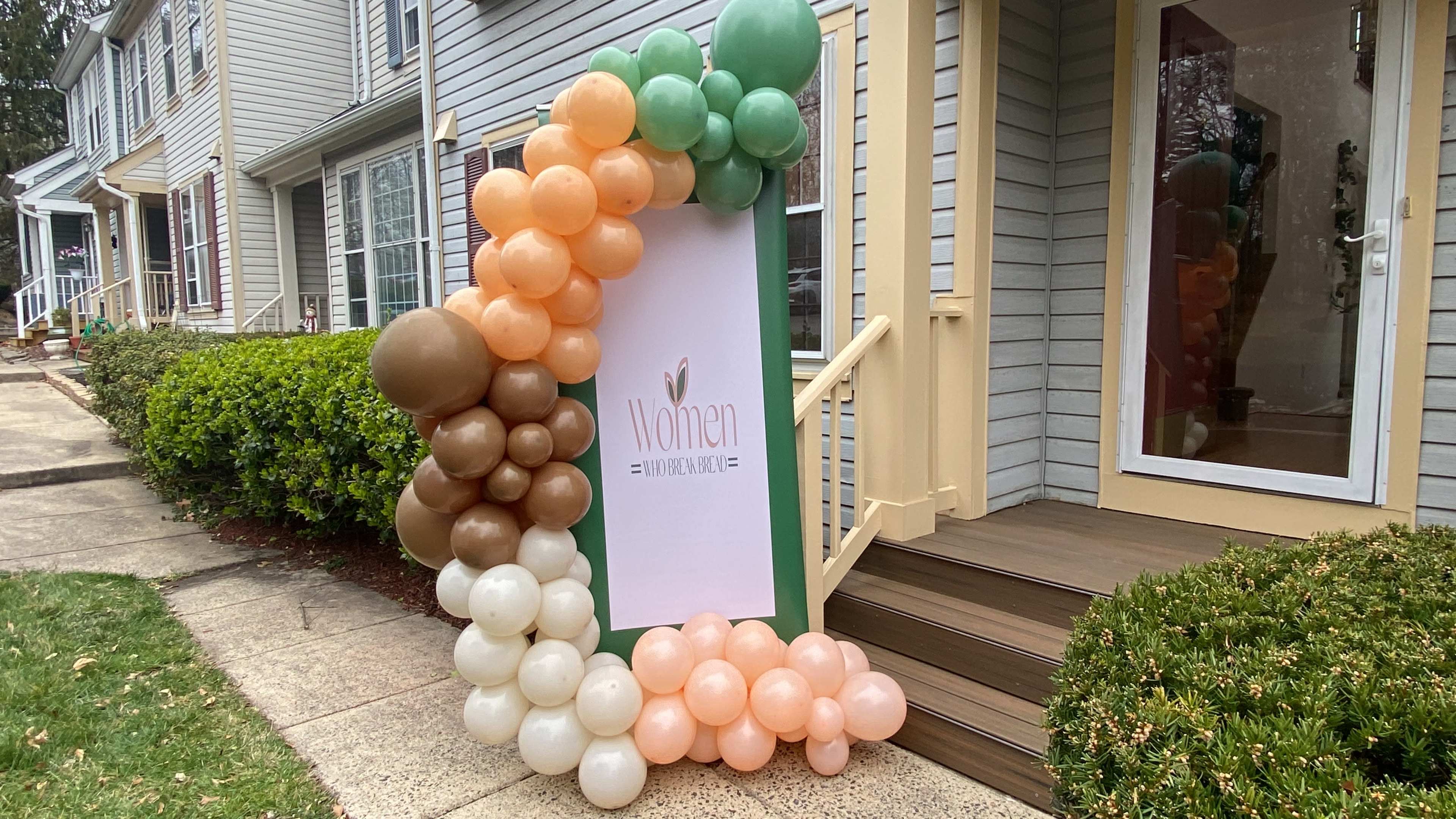 A decorative balloon arch in shades of green, peach, and brown frames a sign that reads "Women" at the entrance of a building.