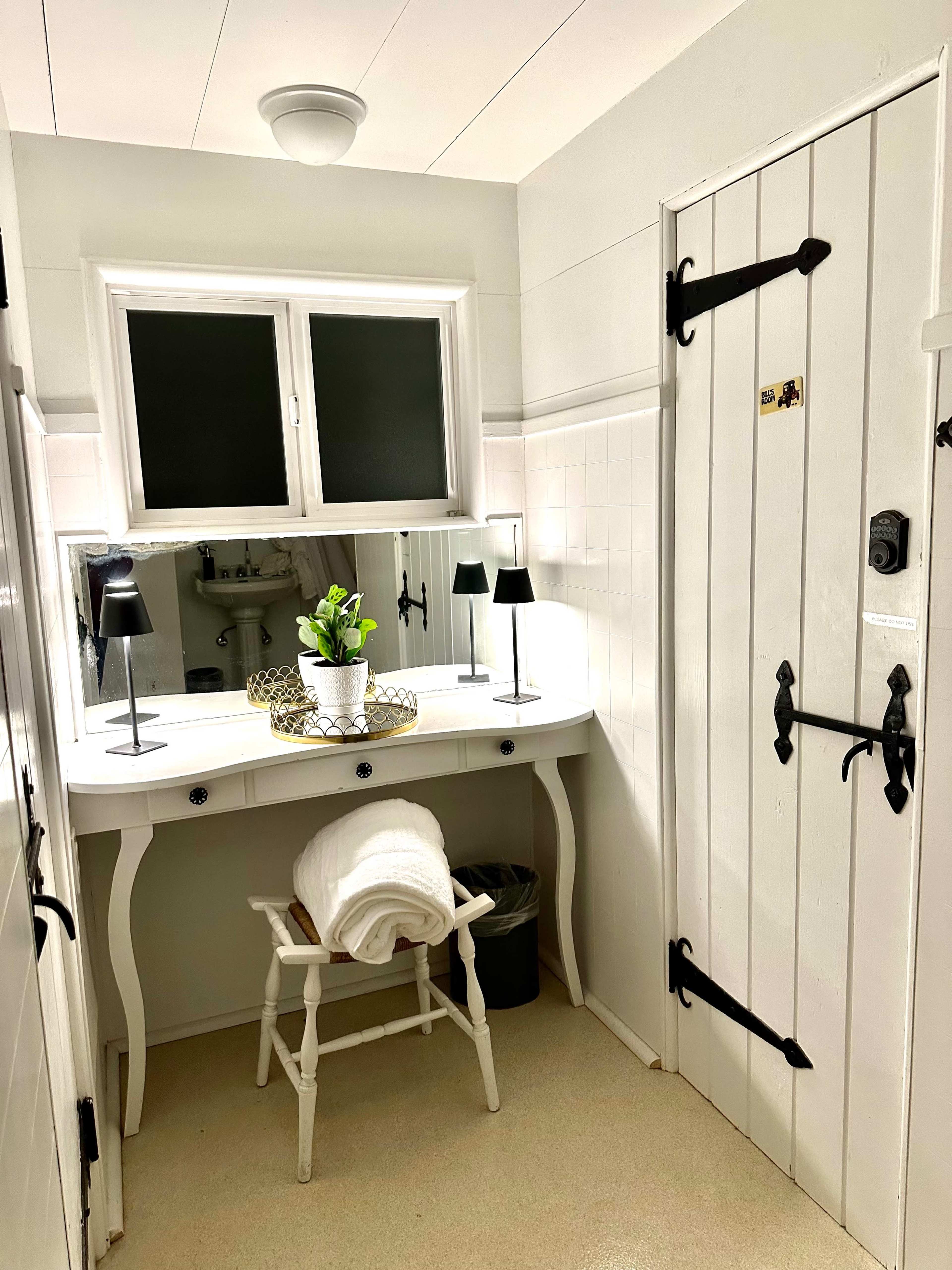 A well-lit bathroom featuring a white vanity with a mirror, two lamps, a potted plant, a towel on a chair, and a paneled door.