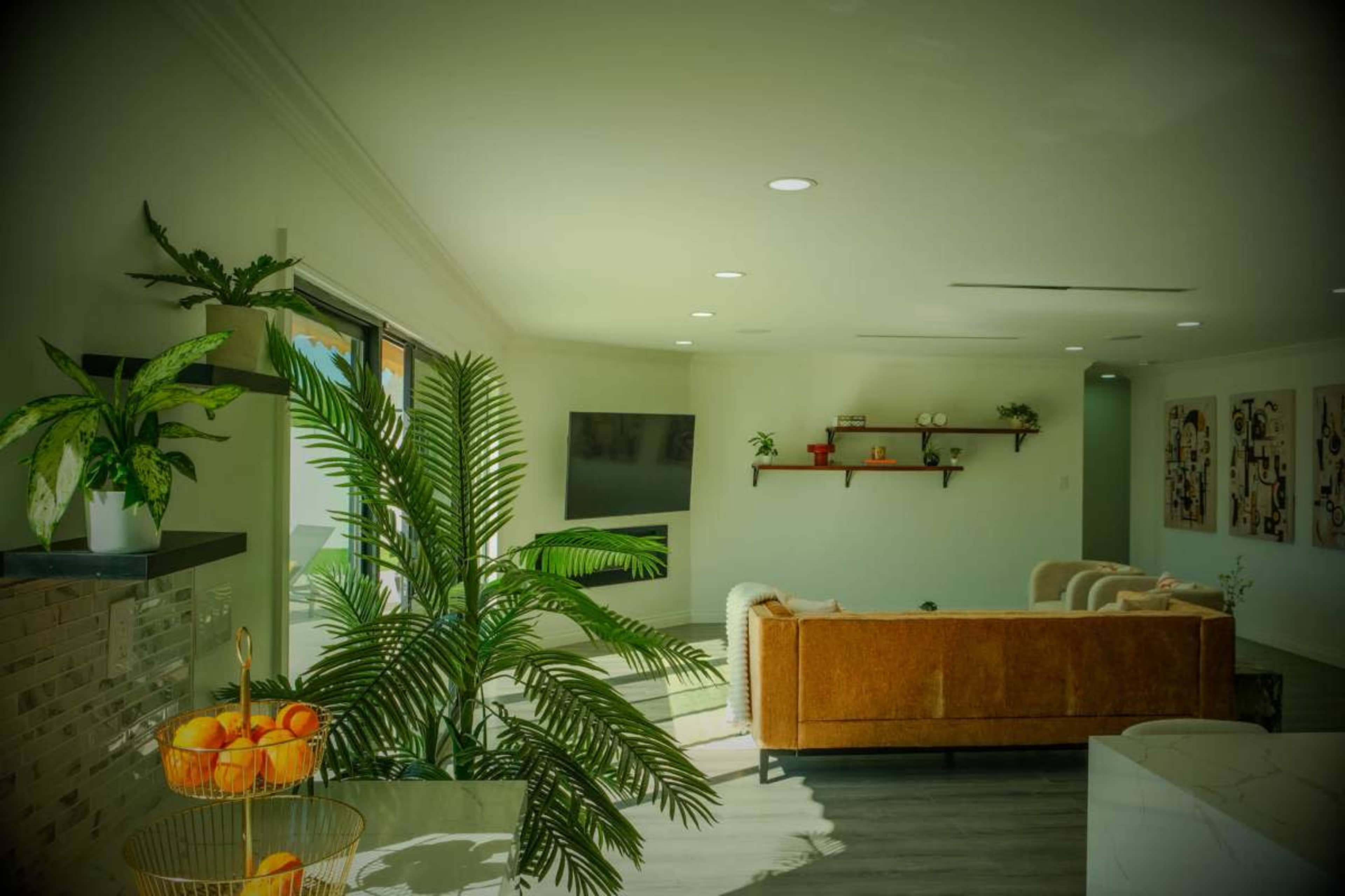 The image shows a modern living room with a large window, a brown sofa, plants, and decorative shelves on the wall.