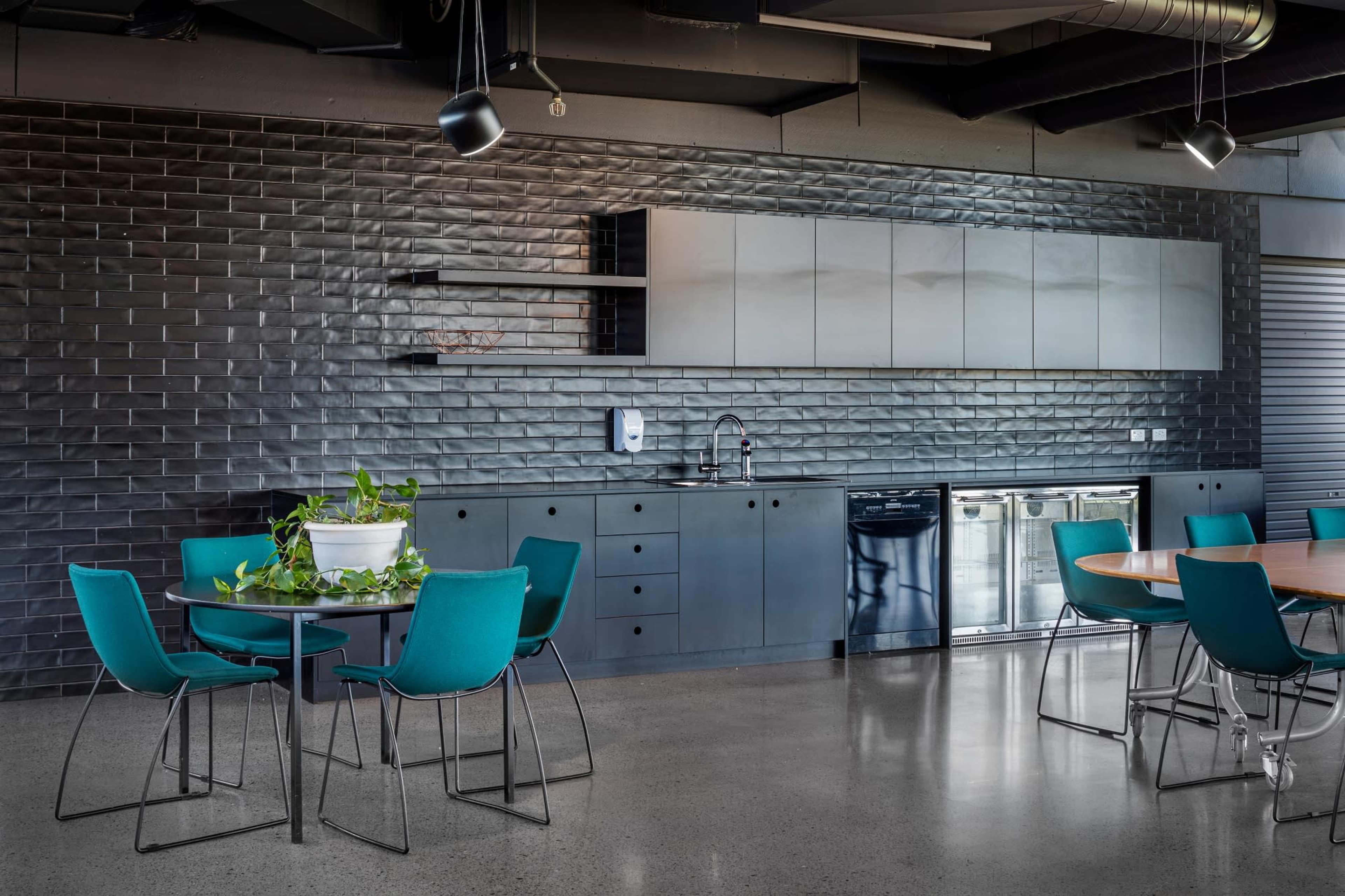 The image shows a modern kitchen with dark tiled walls, metal cabinets, and two dining tables surrounded by teal chairs.