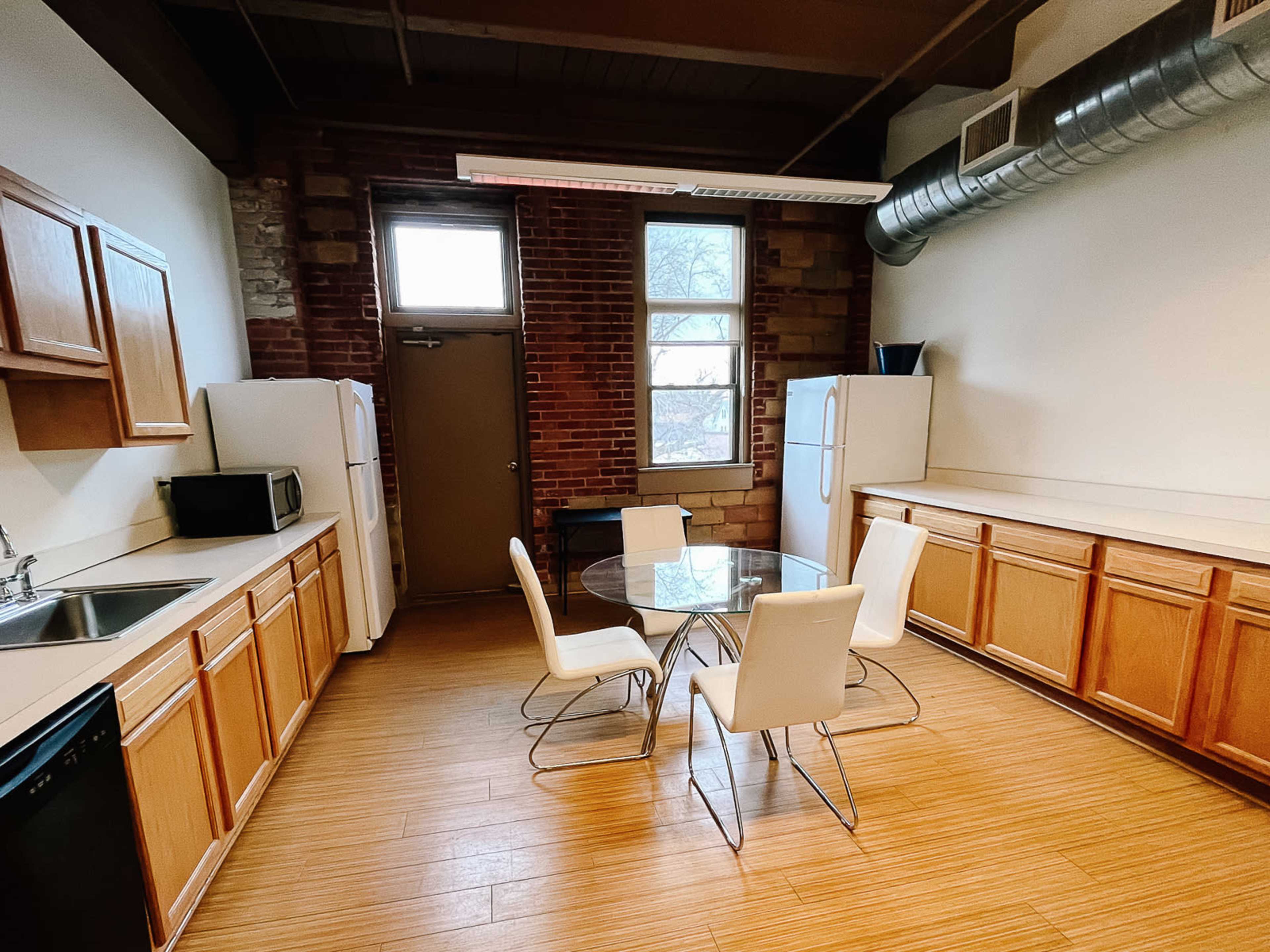 A kitchen with wooden cabinets, a round glass dining table surrounded by four white chairs, and two refrigerators against a brick wall.