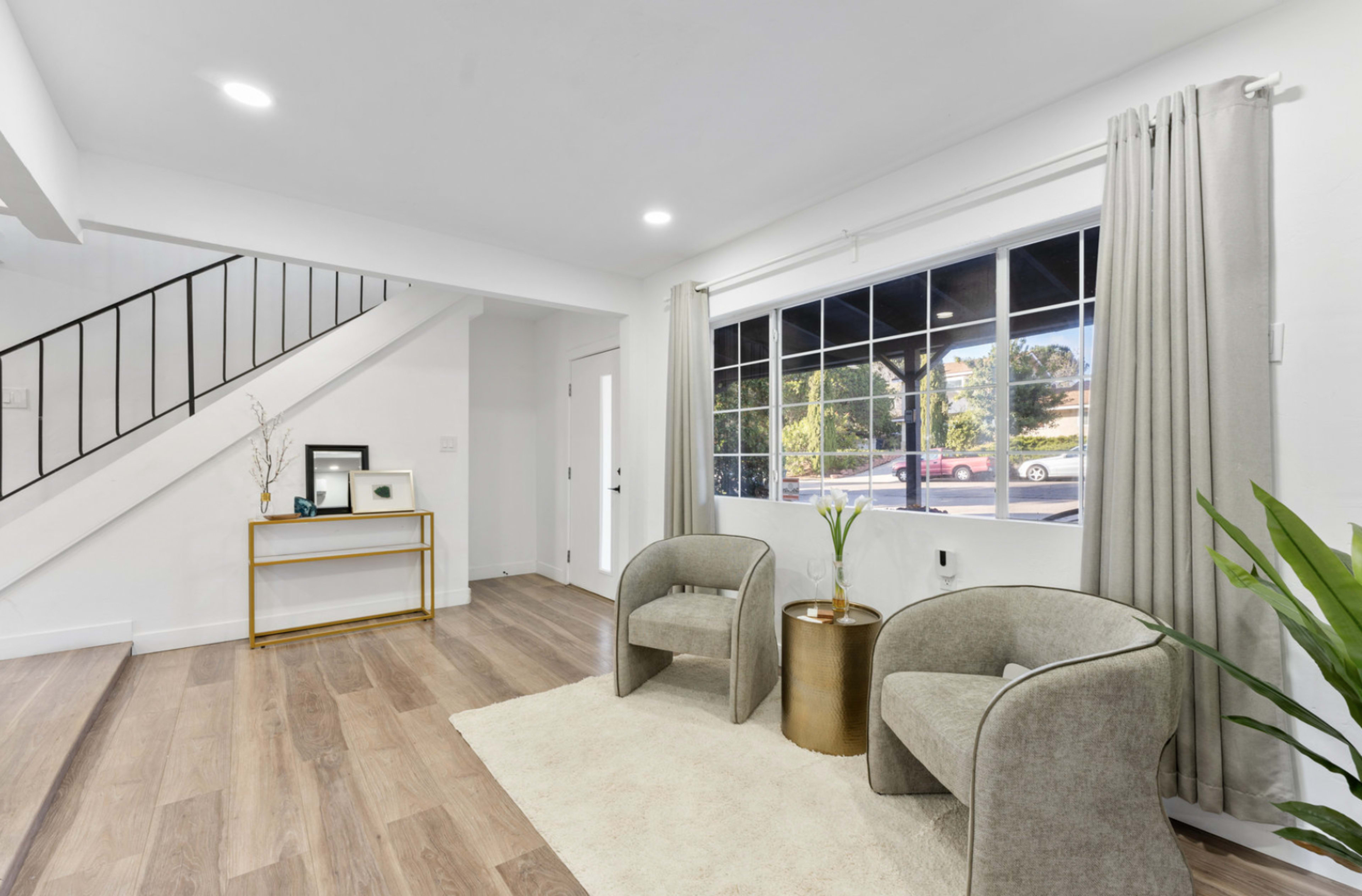 The image shows a living space featuring two armchairs on a rug, a small side table, and a window with curtains, alongside a staircase.