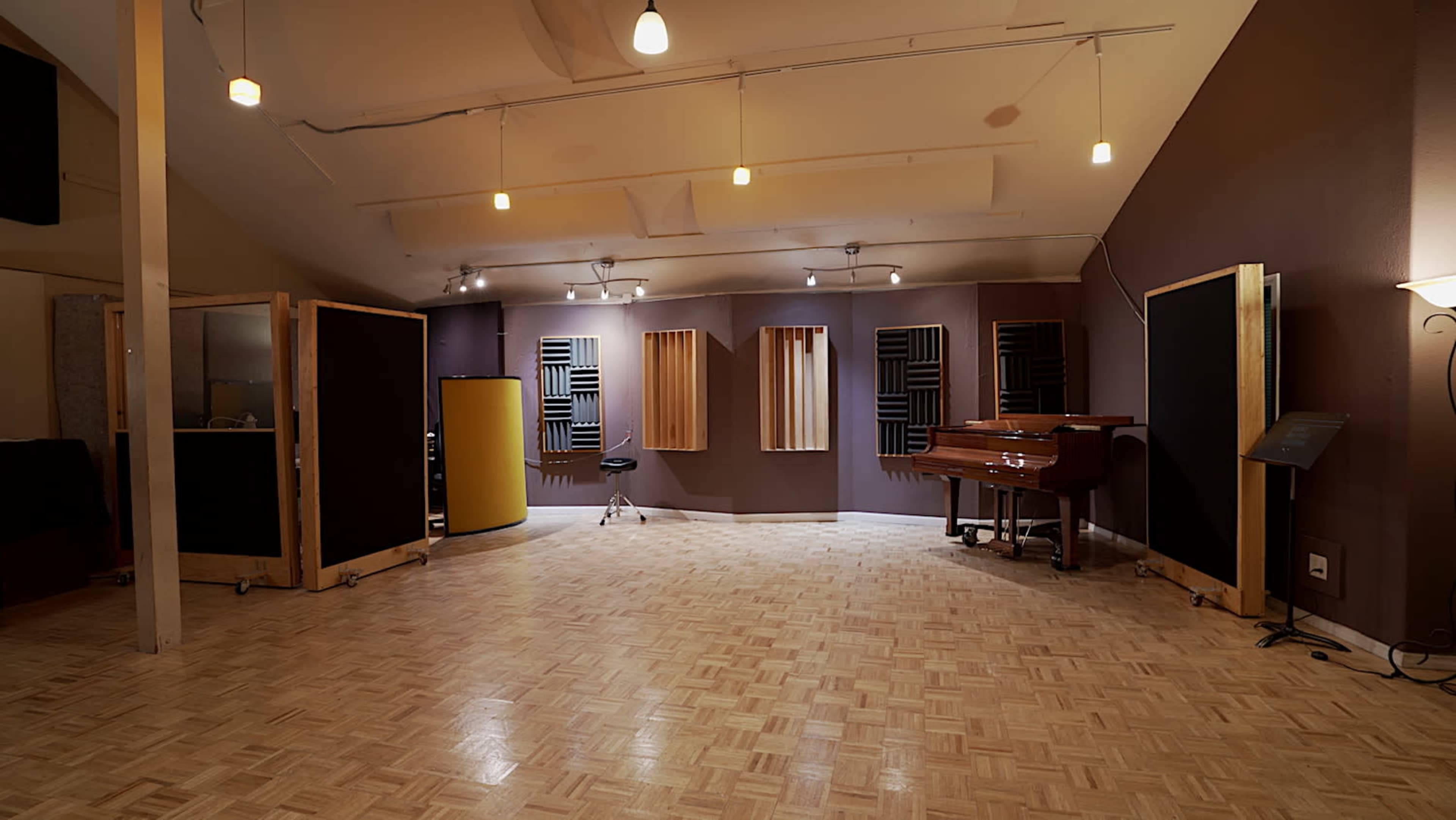 A well-lit recording studio featuring wooden flooring, acoustic panels on the walls, and a grand piano in the center.