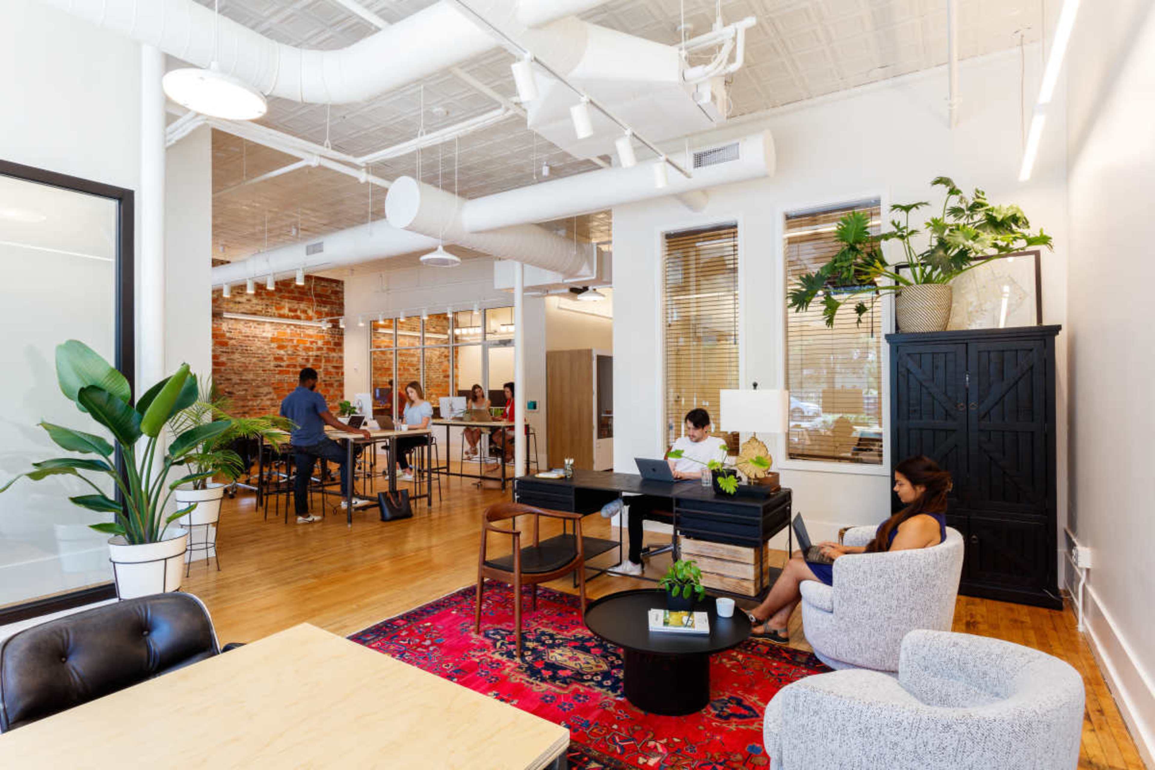 A modern office space features a combination of workstations, a lounge area, and decorative plants, with people engaged in various tasks.