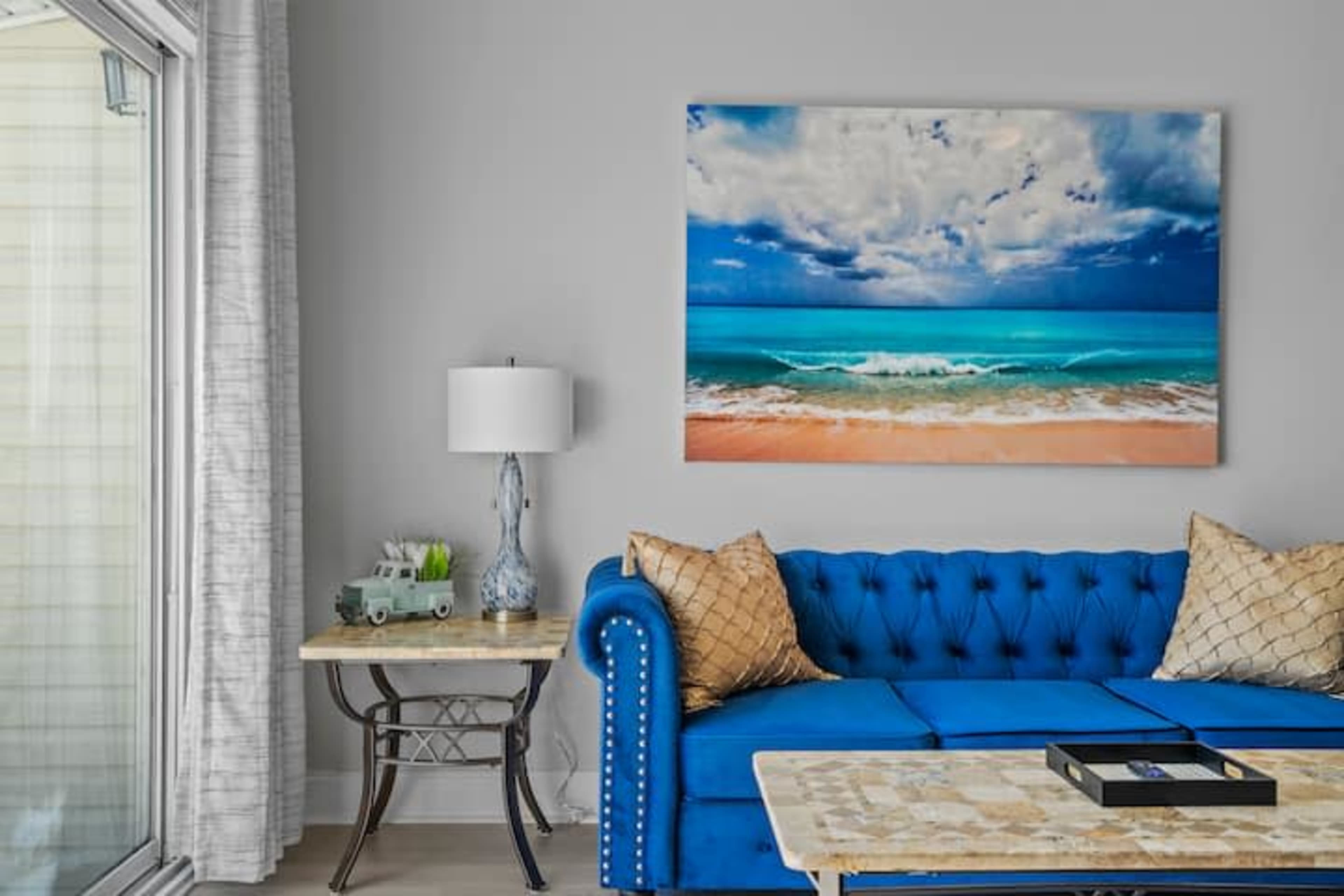 A blue sofa with decorative pillows sits beside a wooden side table and a lamp, in front of a large ocean landscape painting on the wall.