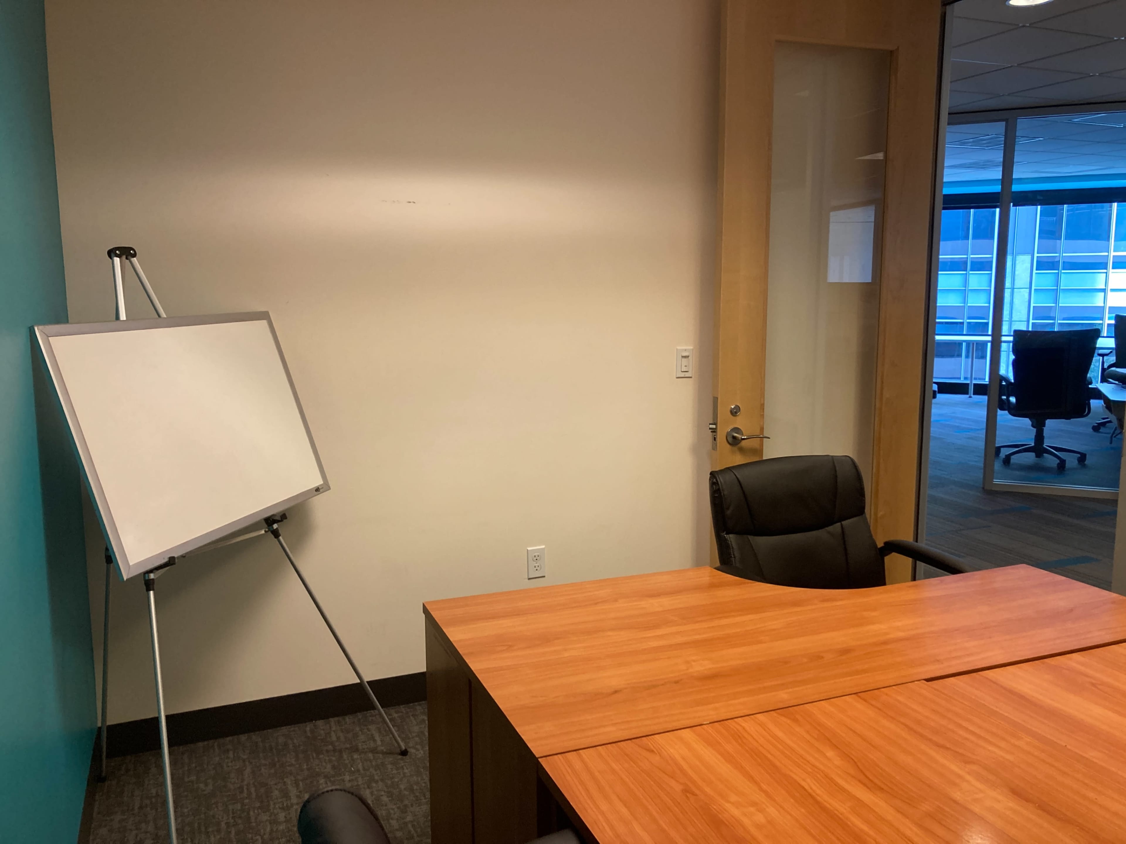 A small office meeting space featuring a wooden table, a black chair, a whiteboard on an easel, and a glass door leading to another area.
