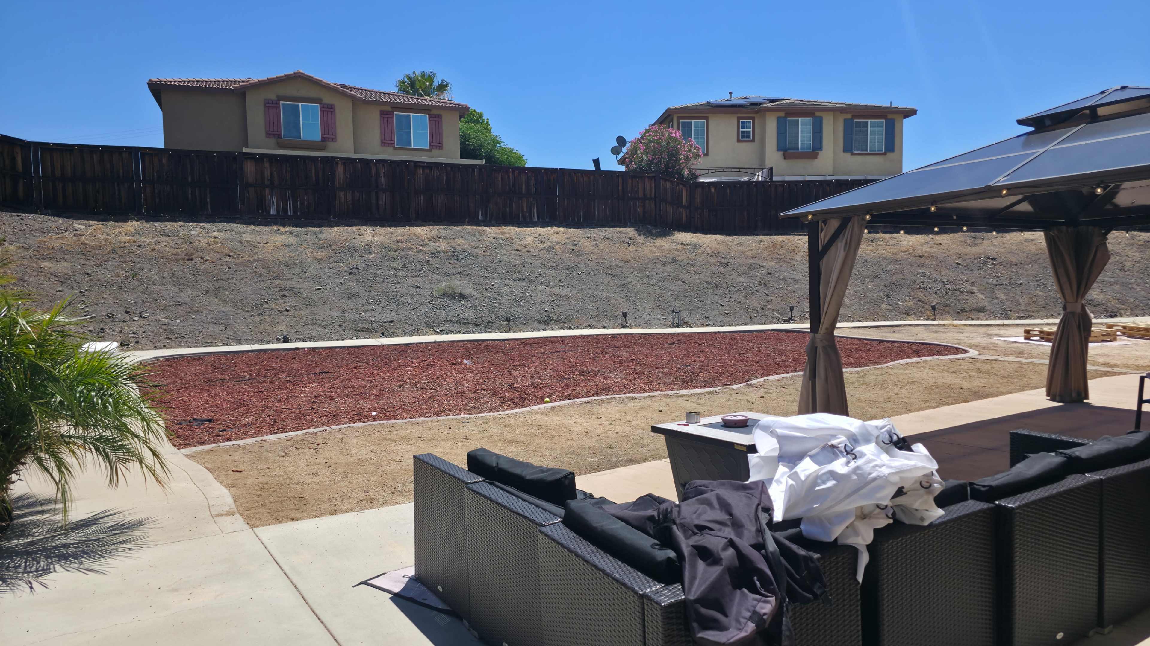 The image shows a dry, landscaped backyard with gravel and a seating area, overlooking a sloped area where houses are positioned above.