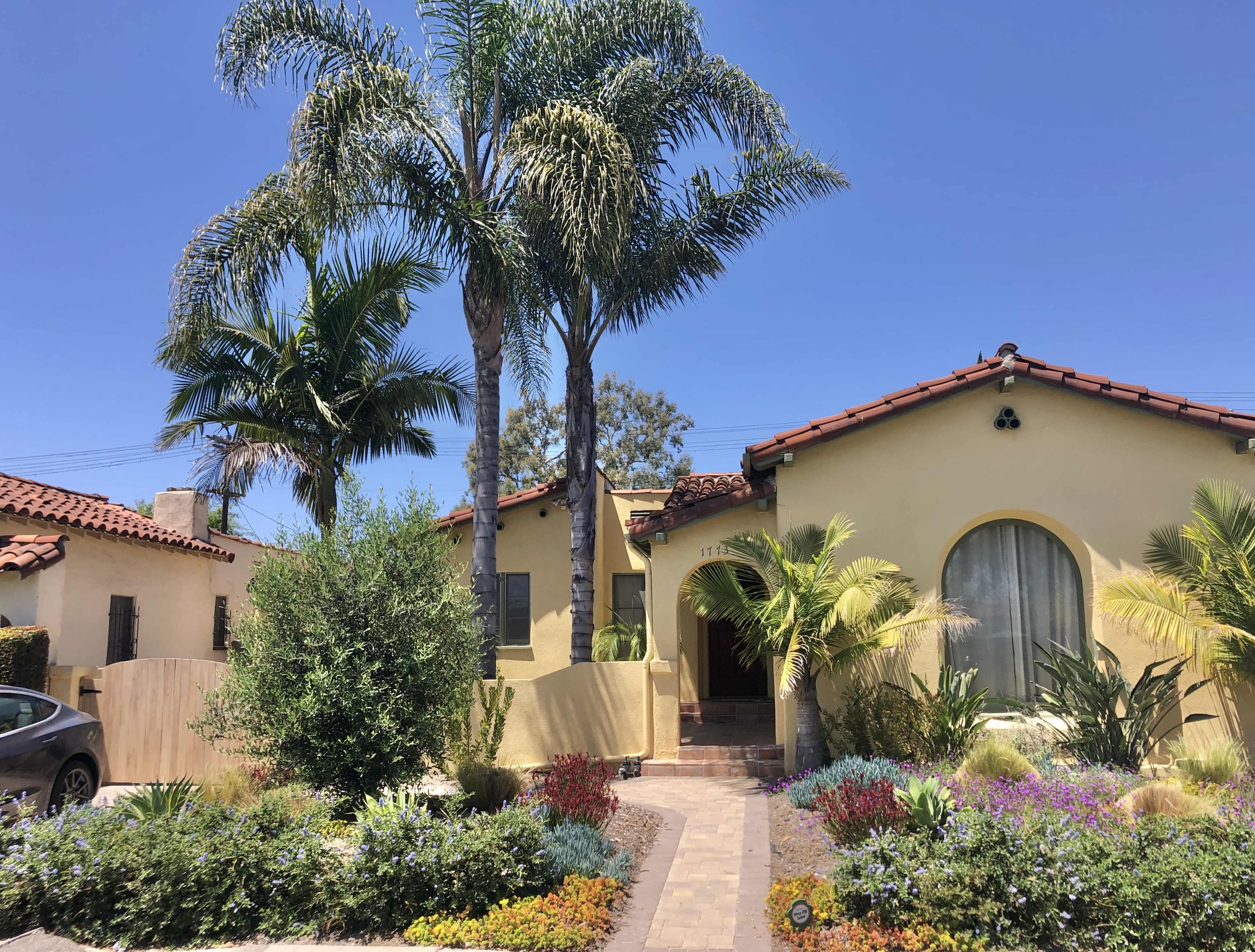 The image shows a yellow stucco house with a red-tile roof, surrounded by palm trees and colorful flower beds in a sunny neighborhood.