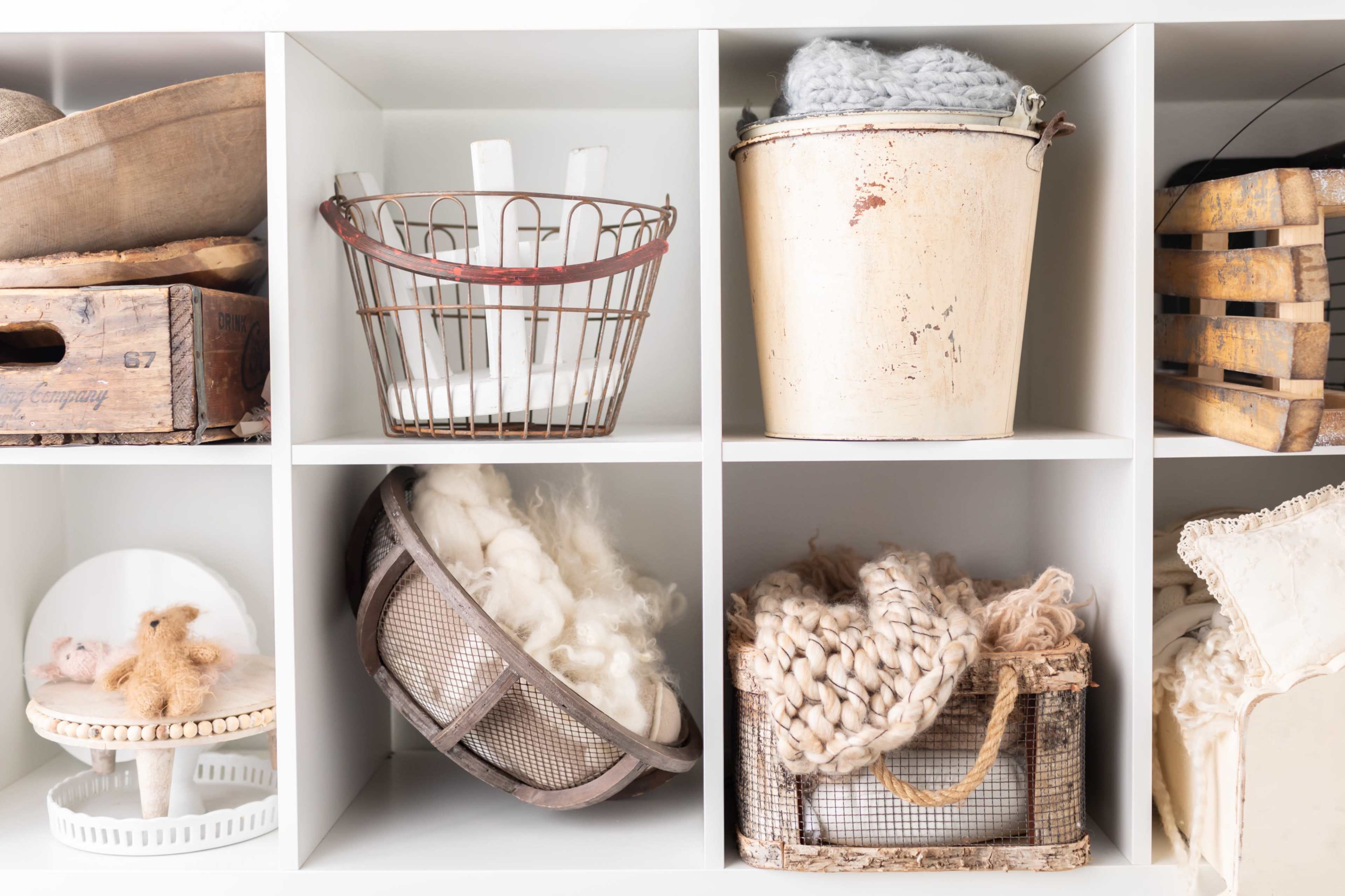 The image shows a white shelving unit filled with various containers, including a metal bucket, a wire basket, and woven storage, alongside wool and textiles.